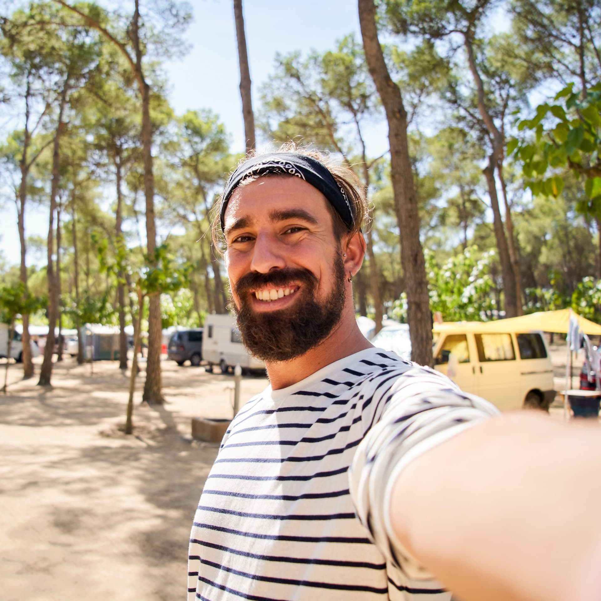 Ein fröhlicher junger Mann mit Vollbart und gestreiftem T-Shirt macht ein Selfie auf einem Campingplatz mit Wohnmobilen in einem Kiefernwald.