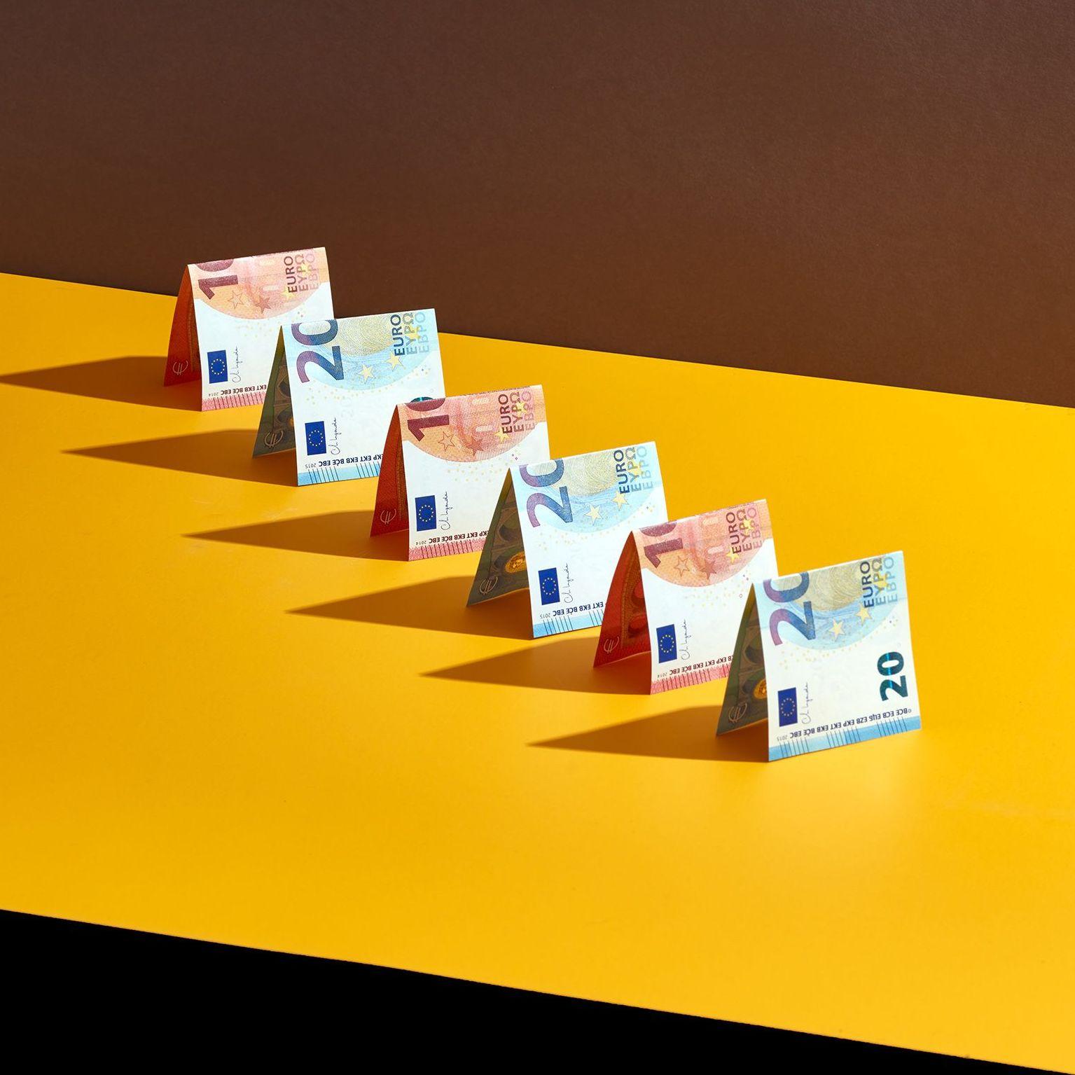 Mehrere Geldscheine stehen wie Häuser gefaltet in einer Reihe auf gelbem Untergrund. 
