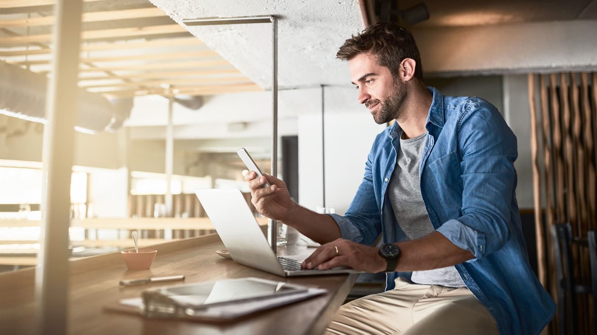 Junger bärtiger Mann arbeitet mit Laptop, Tablet und Handy im Cafe.