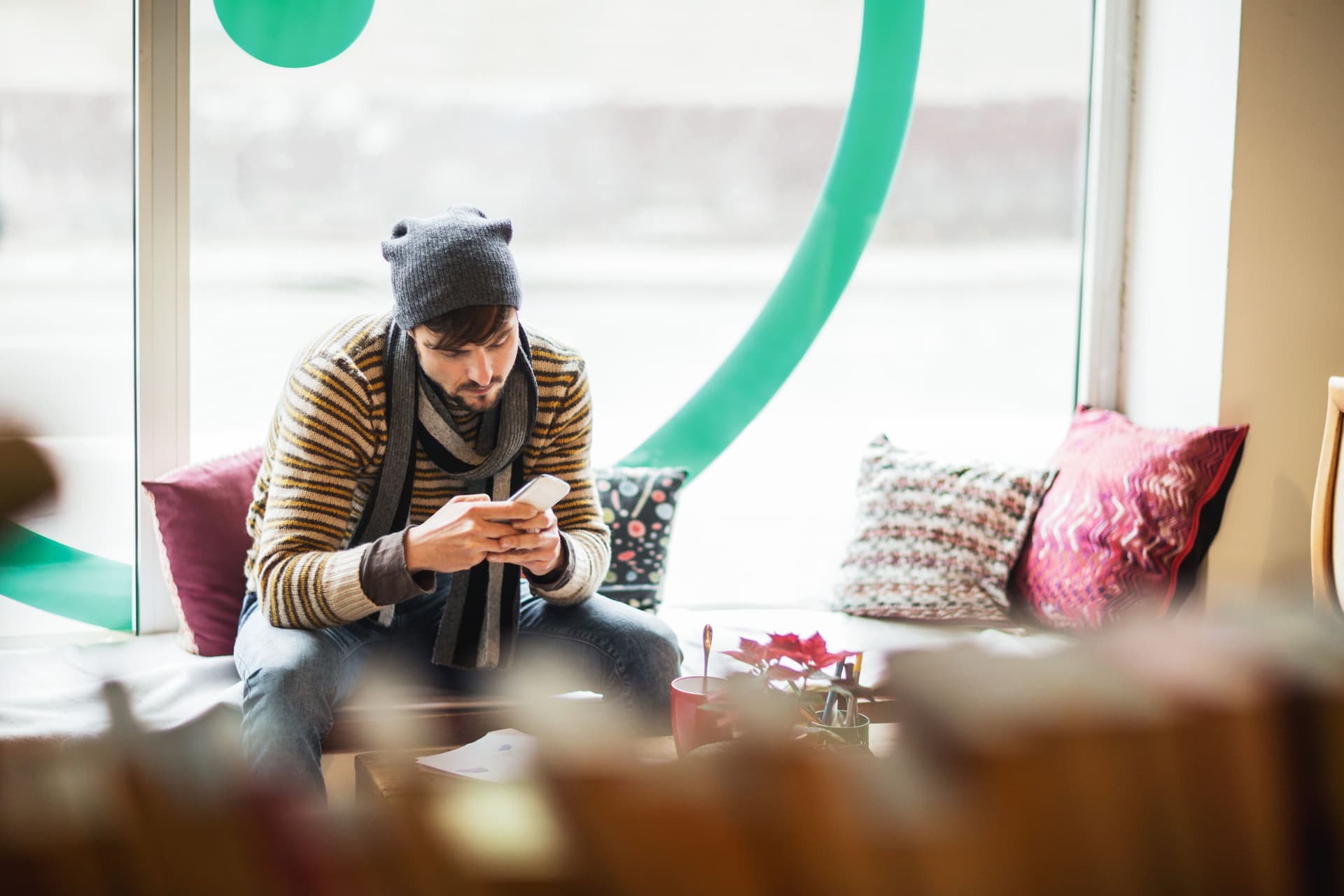 Junger Mann mit Mütze und Schal im Cafe schaut auf sein Handy.