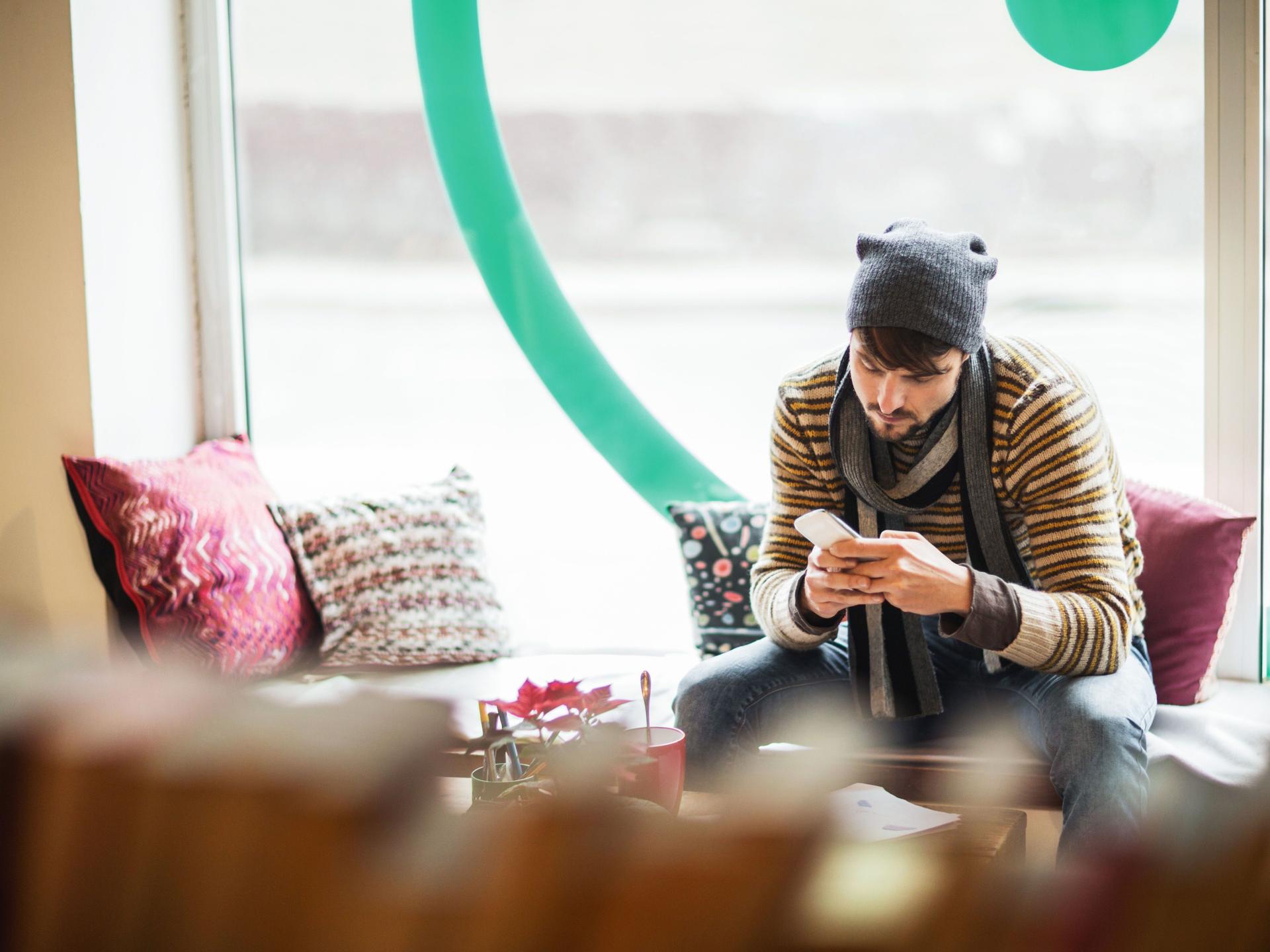 Junger Mann mit Mütze und Schal im Cafe schaut auf sein Handy.
