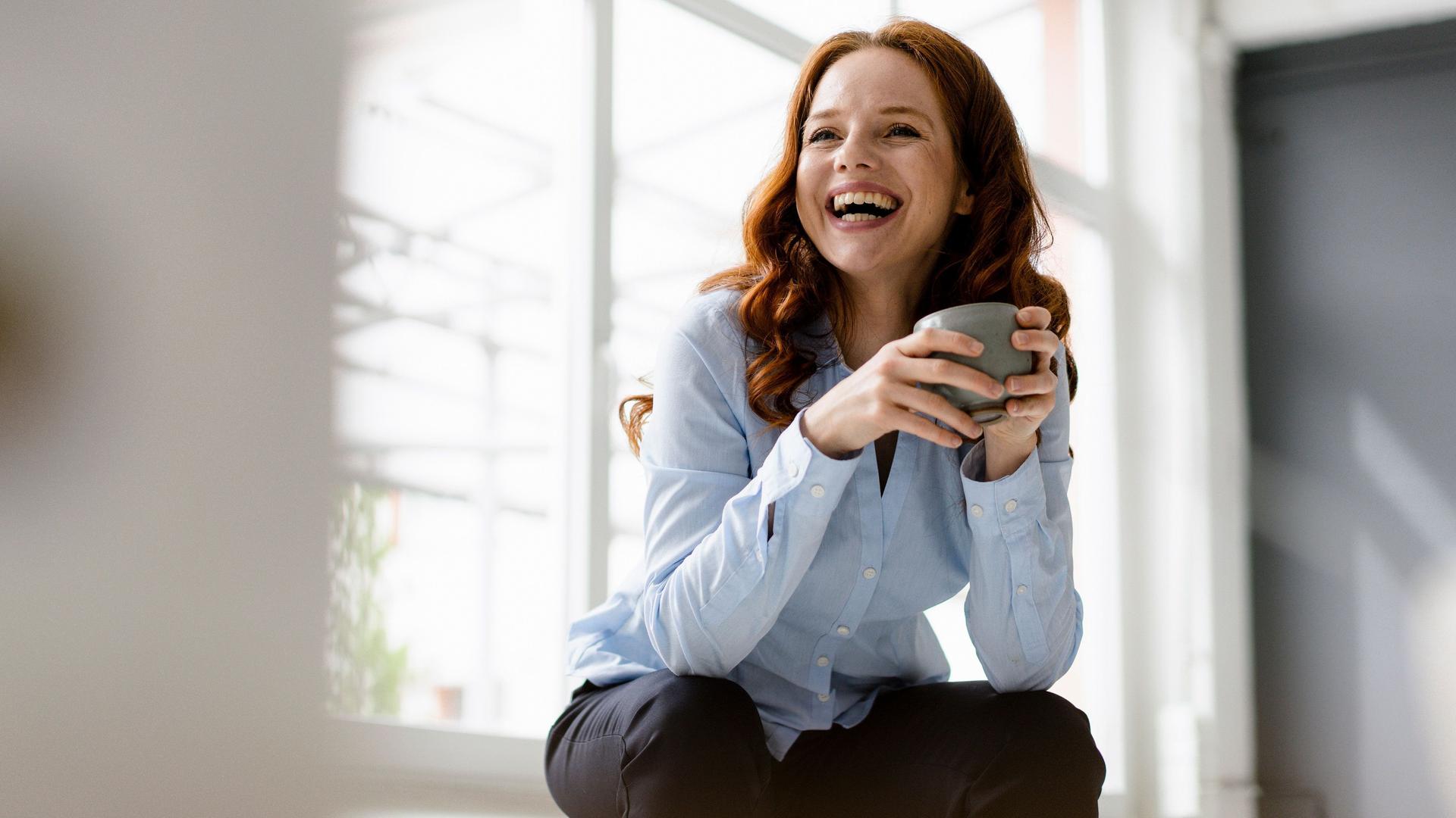 Portrait of laughing redheaded woman with tea bowl sitting on backrest in a loft
