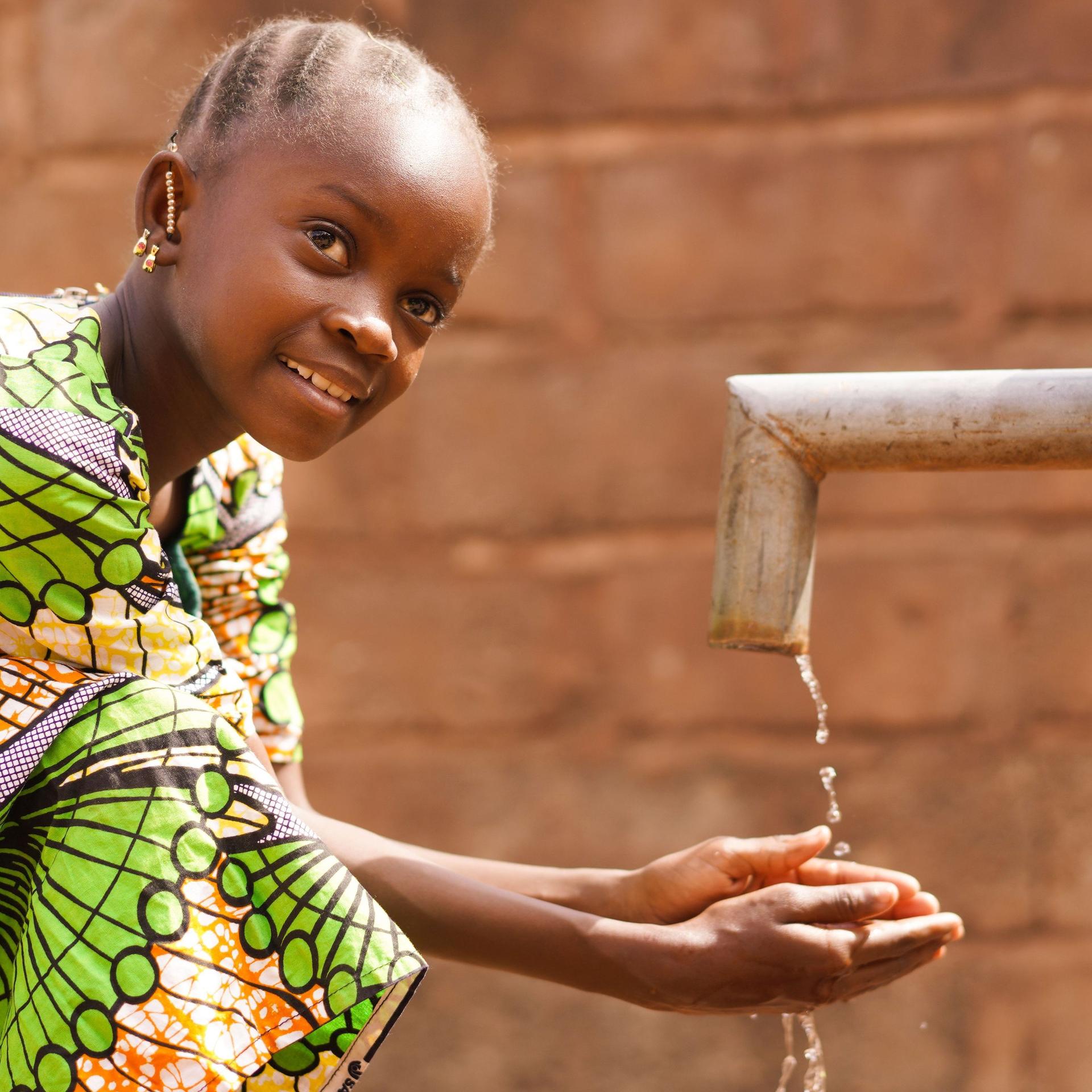 Kind in einem afrikanischen Land wäscht sich an einem Brunnen die Hände und lächelt in die Kamera.