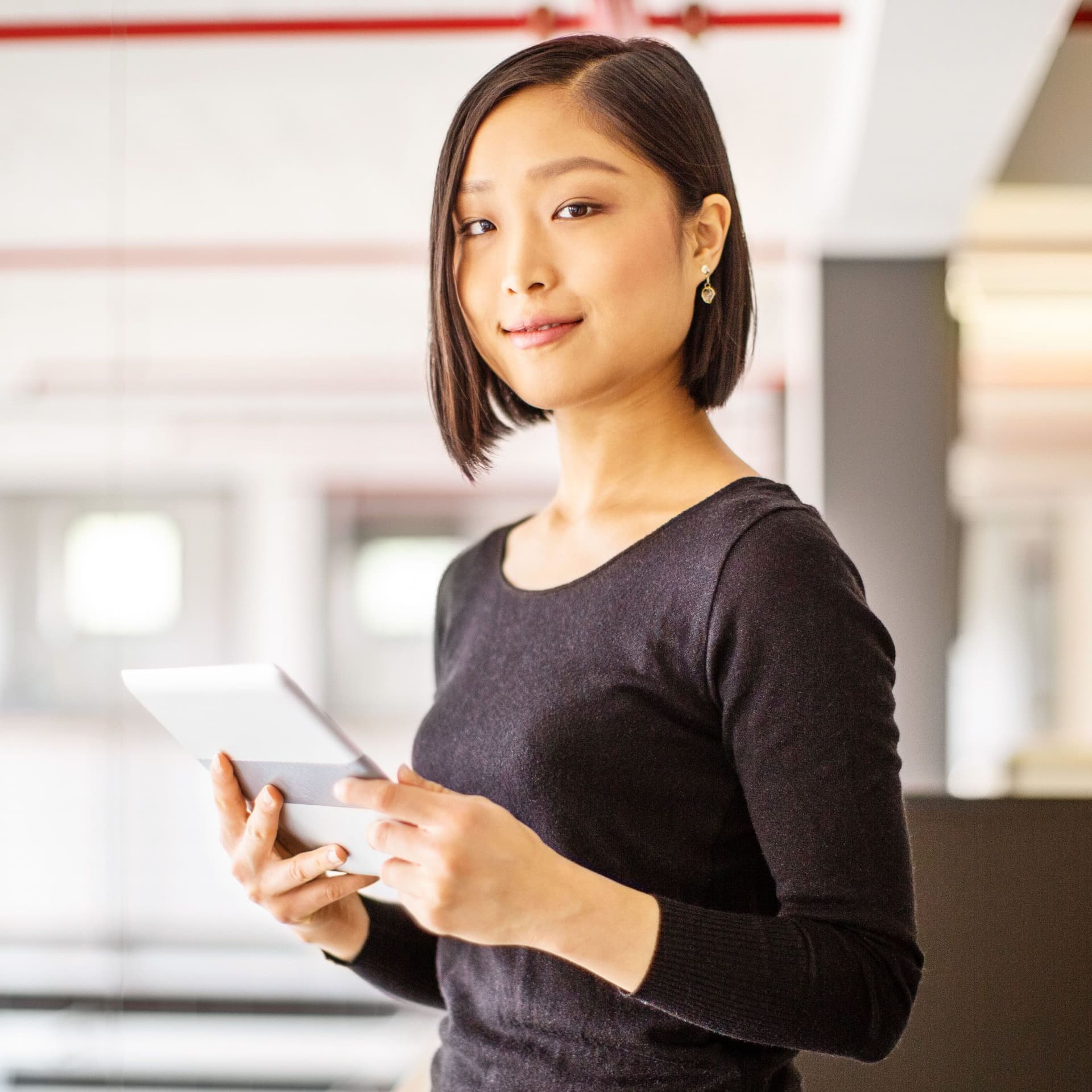 Eine junge Frau hält ein silbernes Tablet in der Hand, sie blickt seitlich in die Kamera. Im Hintergrund sieht man Tische und Computer in einem Büro.