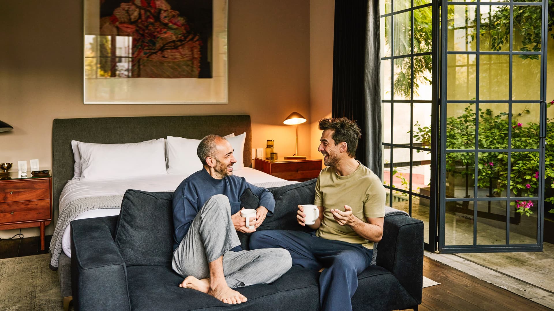 Zwei Männer sitzen entspannt auf einem Sofa in einem gemütlichen Schlafzimmer, trinken Kaffee und unterhalten sich lachend, während durch die geöffnete Glastür ein grüner Garten zu sehen ist.