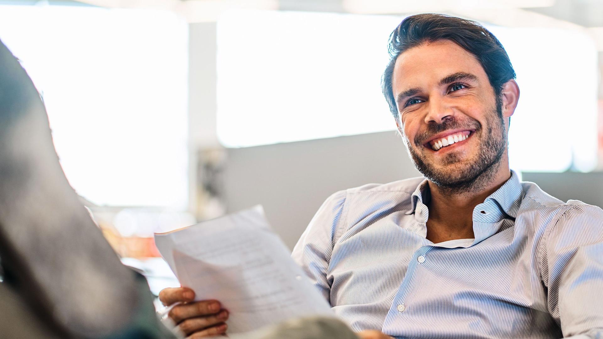 Lächelnder junger Mann, der Papiere liest, während er mit den Füßen hoch auf seinem Schreibtisch in einem großen, modernen Büro sitzt.