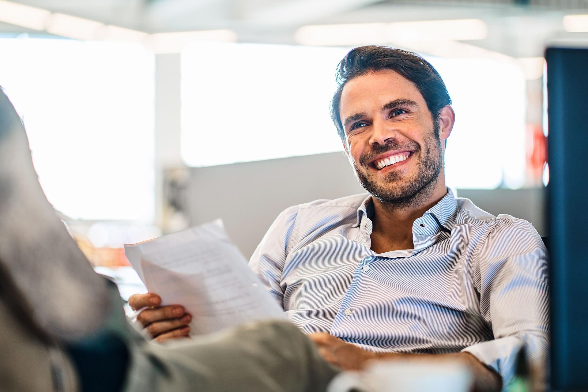Lächelnder junger Mann, der Papiere liest, während er mit den Füßen hoch auf seinem Schreibtisch in einem großen, modernen Büro sitzt.