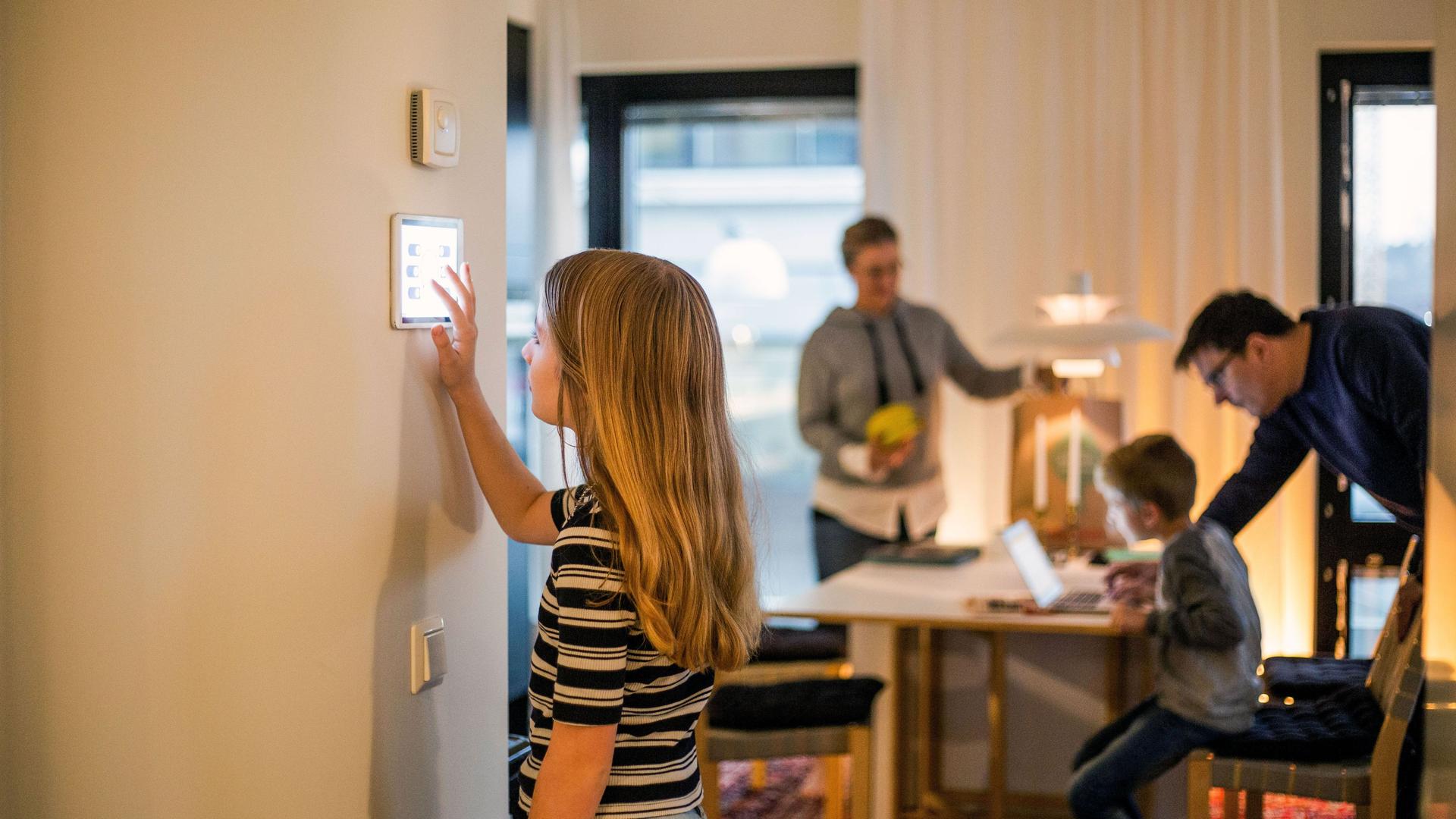 Mädchen bedient digitales Smart Home Display an der Wand. Im Hintergrund ist unscharf die Familie zu erkennen.