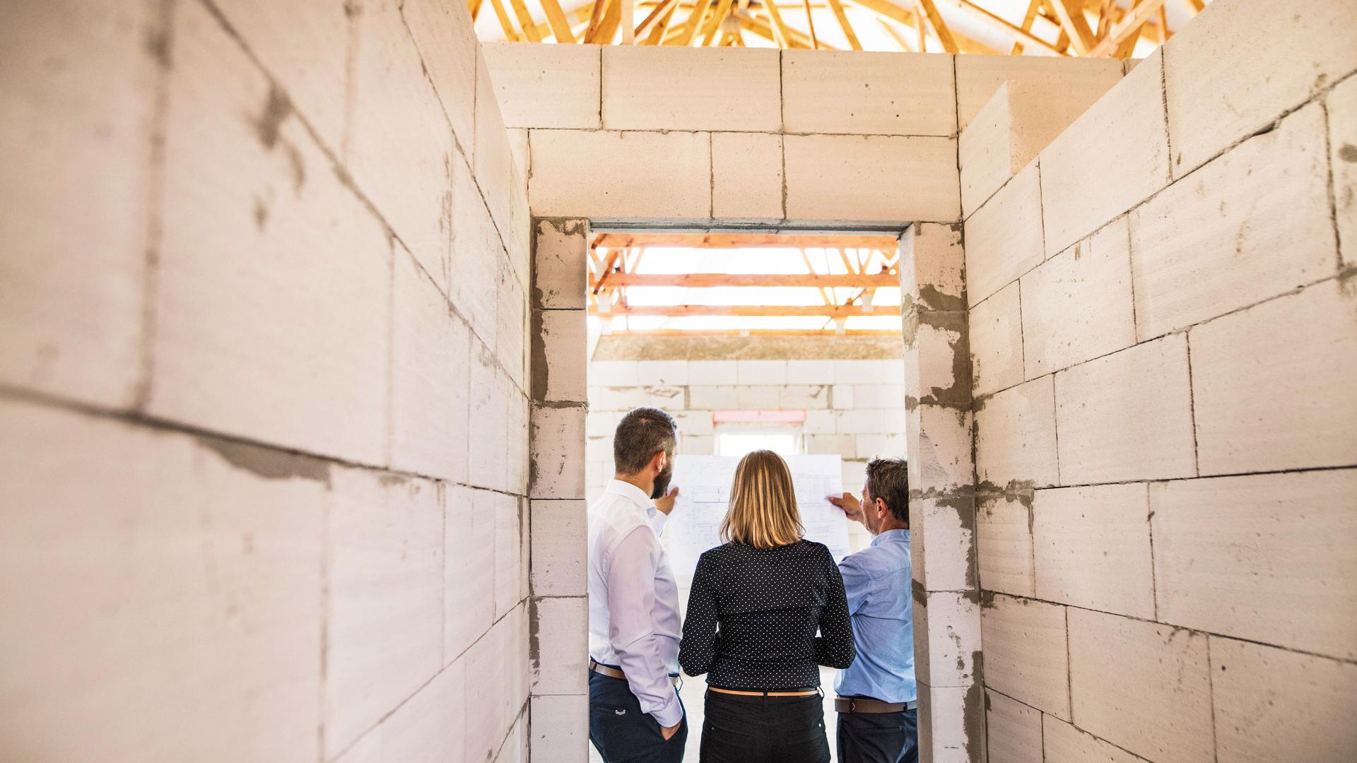 Drei Personen stehen mit dem Rücken zur Kamera auf der Baustelle eines Neubaus.