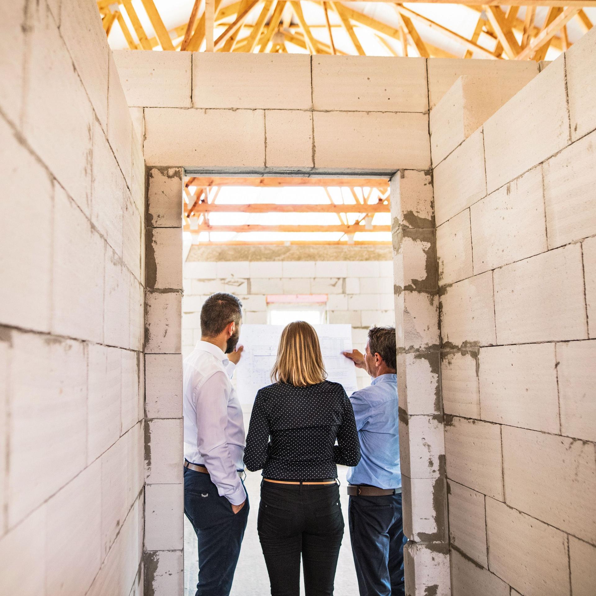 Drei Personen stehen mit dem Rücken zur Kamera auf der Baustelle eines Neubaus.