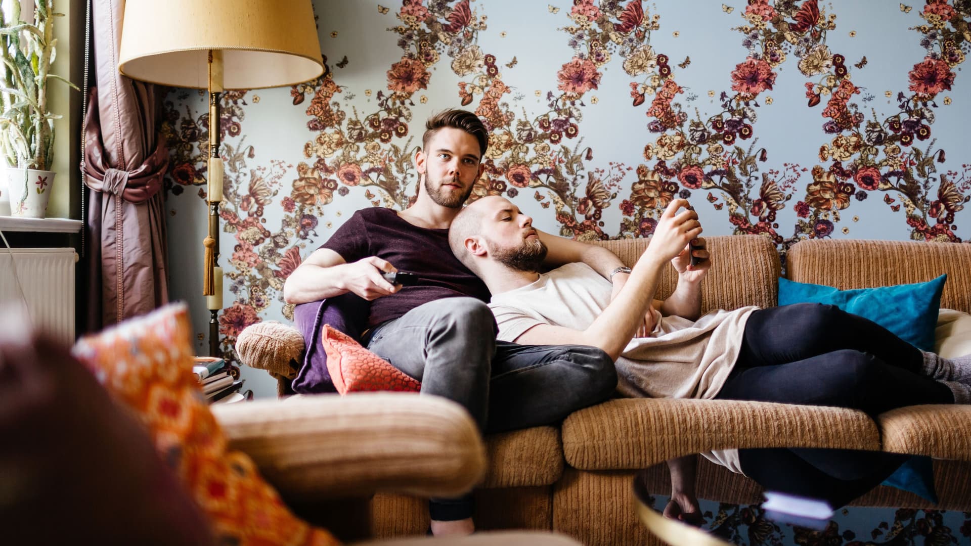 Zwei Männer liegen zusammen auf dem Sofa. Ein Mann schaut in sein Smartphone, der andere hält einen Fernsehdrücker in der Hand.