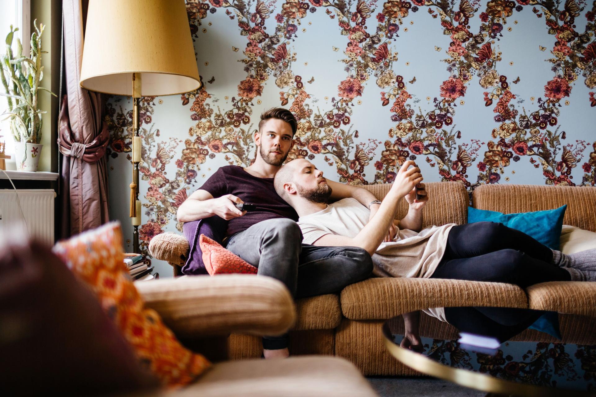 Zwei Männer liegen zusammen auf dem Sofa. Ein Mann schaut in sein Smartphone, der andere hält einen Fernsehdrücker in der Hand.