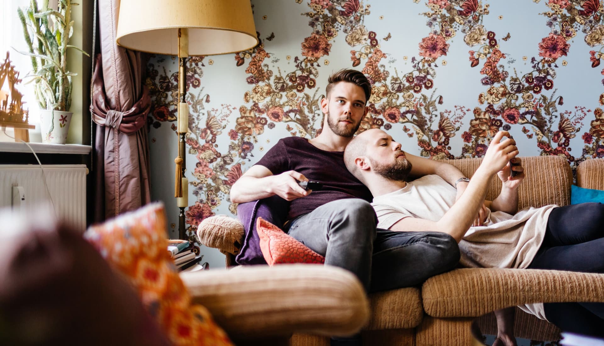 Zwei Männer liegen zusammen auf dem Sofa. Ein Mann schaut in sein Smartphone, der andere hält einen Fernsehdrücker in der Hand.