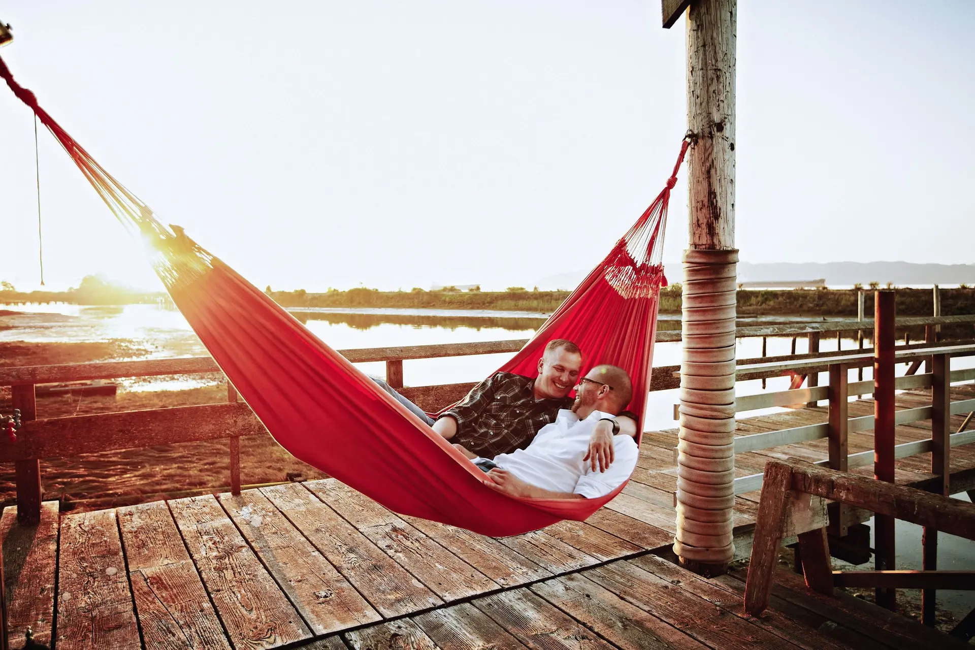 Ein schwules Paar liegt in einer Hängematte am Dock bei Sonnenuntergang. Sie umarmen sich und lachen.