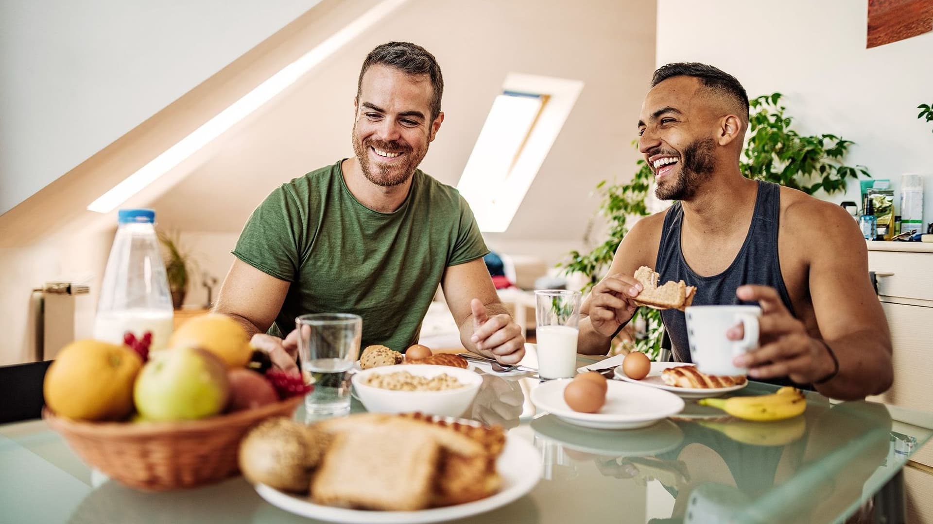 Zwei Männer frühstücken ausgelassen miteinander. Sie sitzen der Kamera zugewandt und vor ihnen ist ein gedeckter Tisch mit gesunden Nahrungsmitteln. Im Hintergrund sieht man die Schrägen einer Dachgeschosswohnung.