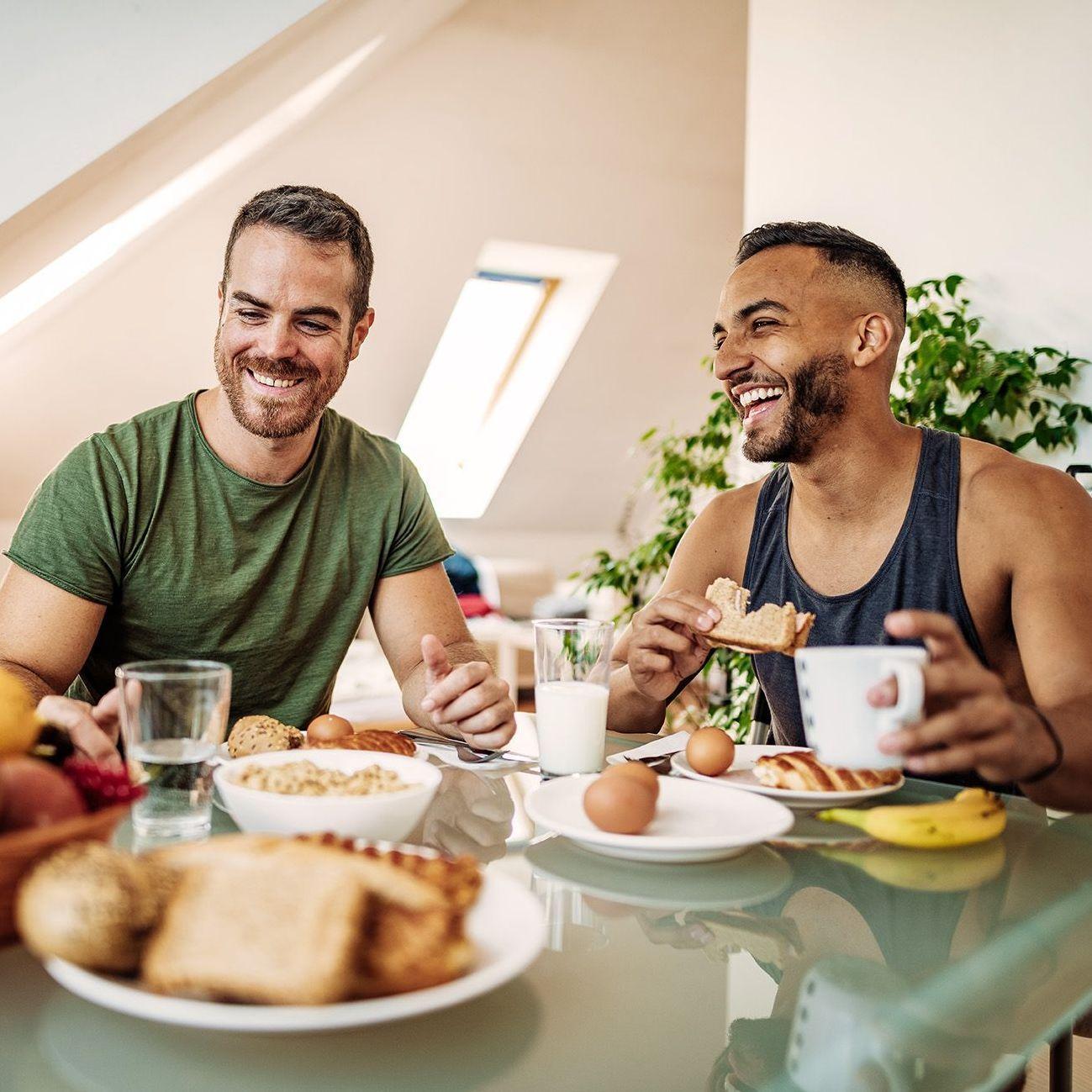 Zwei Männer frühstücken ausgelassen miteinander. Sie sitzen der Kamera zugewandt und vor ihnen ist ein gedeckter Tisch mit gesunden Nahrungsmitteln. Im Hintergrund sieht man die Schrägen einer Dachgeschosswohnung.