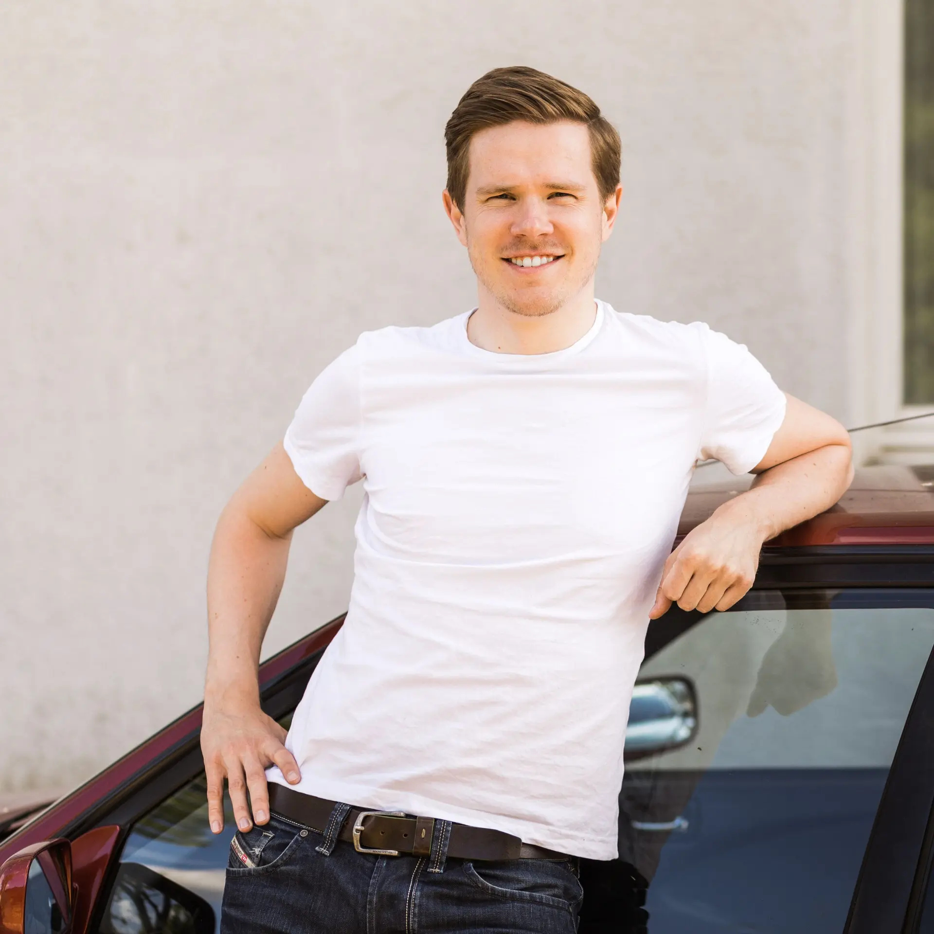 Ein junger Mann steht angelehnt an einem Auto und lächelt.