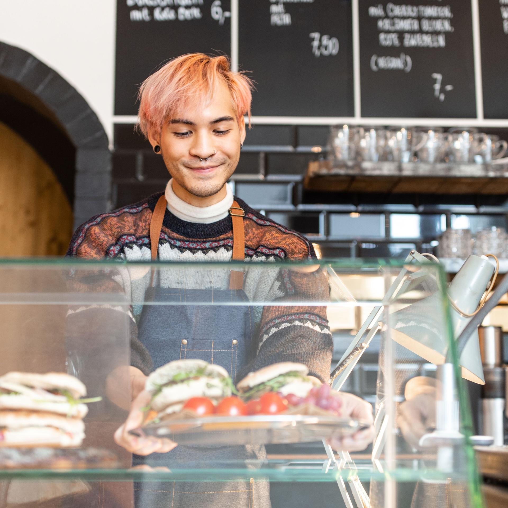 Ein Mann mit rosa gefärbten Haaren arbeitet in einem Cafe und befüllt die Theke mit Paninis.