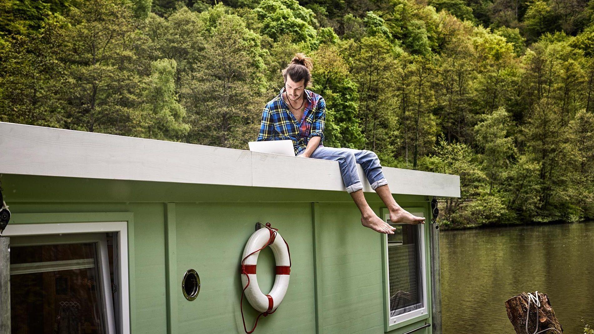 Man mit man bun sitzt barfuss mit einem Laptop auf dem Dach eines Hausboots.