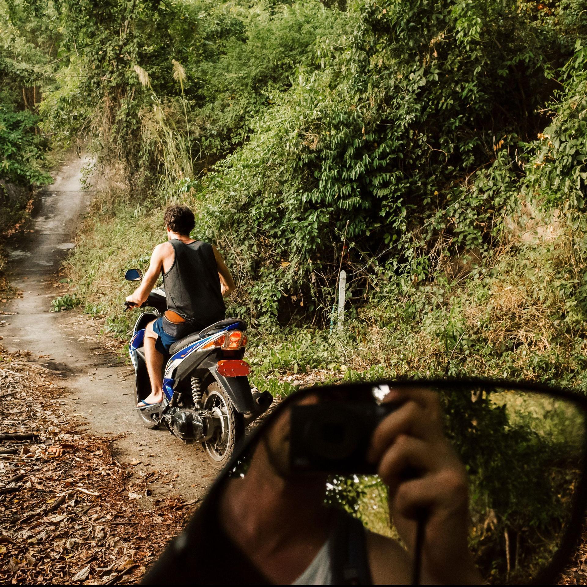 Junger Mann fährt auf Scooter einen Dschungelpfad entlang. Ein anderer Mann spiegelt sich im Rückspiegel während er ein Foto macht.