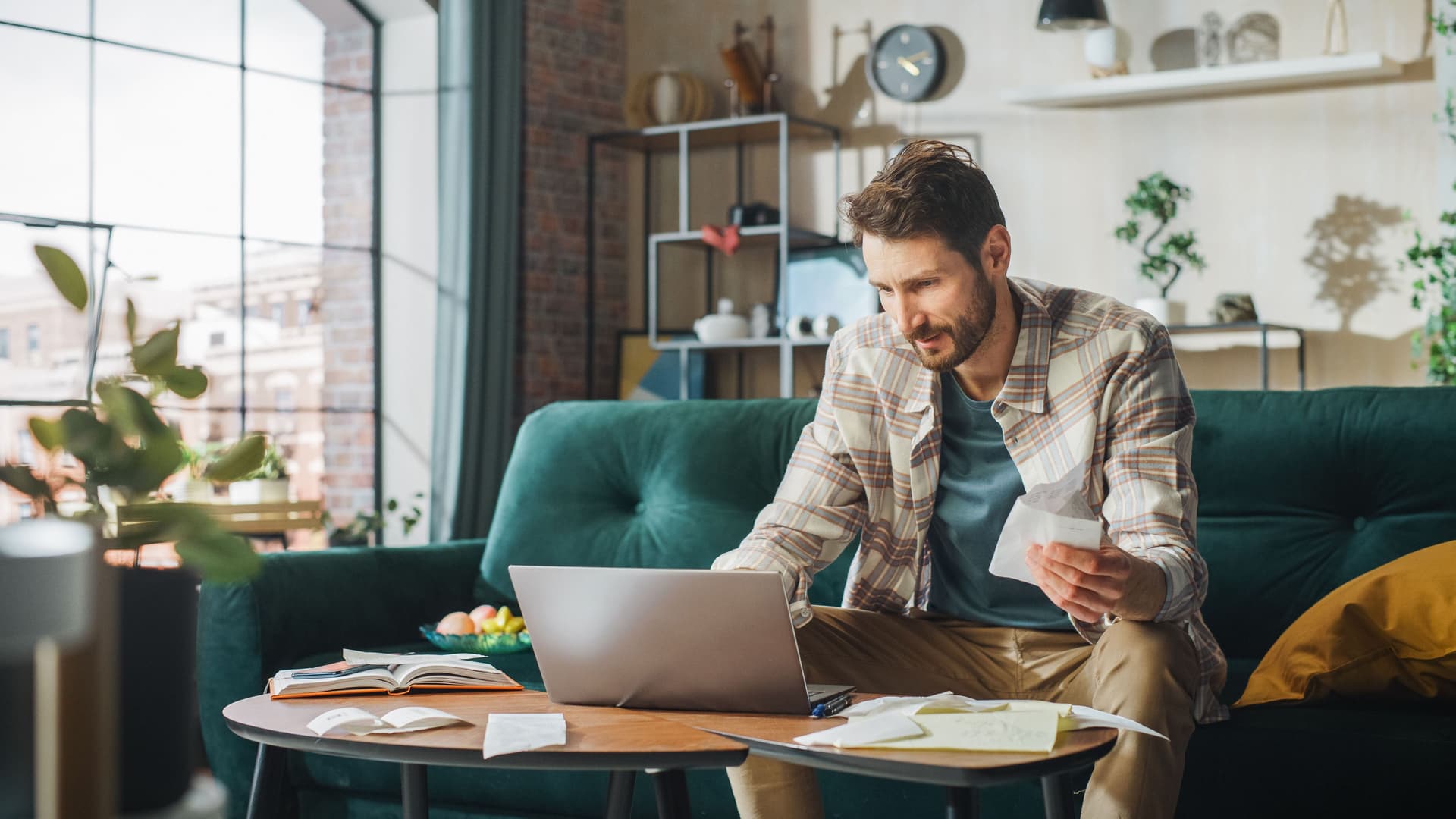 Ein Mann in den dreißigern bezahlt Rechnungen im Online-Banking.Er sitzt auf einer smaragdfarbenen Samt-Couch.