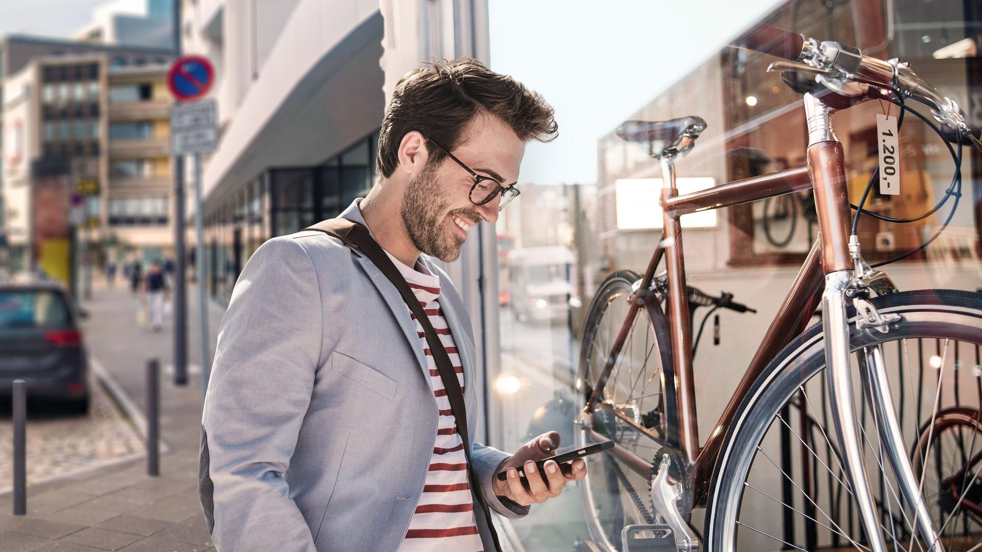 Mann mit Blick auf sein Smartphone, steht auf einer Straße vor einem Fahrradladen.