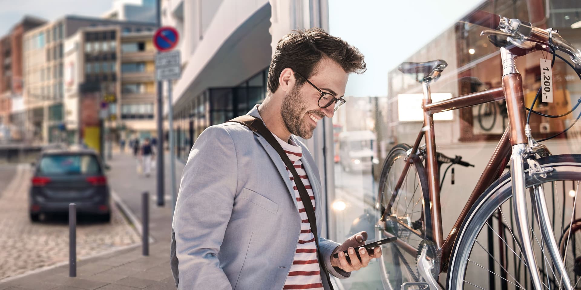 Mann mit Blick auf sein Smartphone, steht auf einer Straße vor einem Fahrradladen.