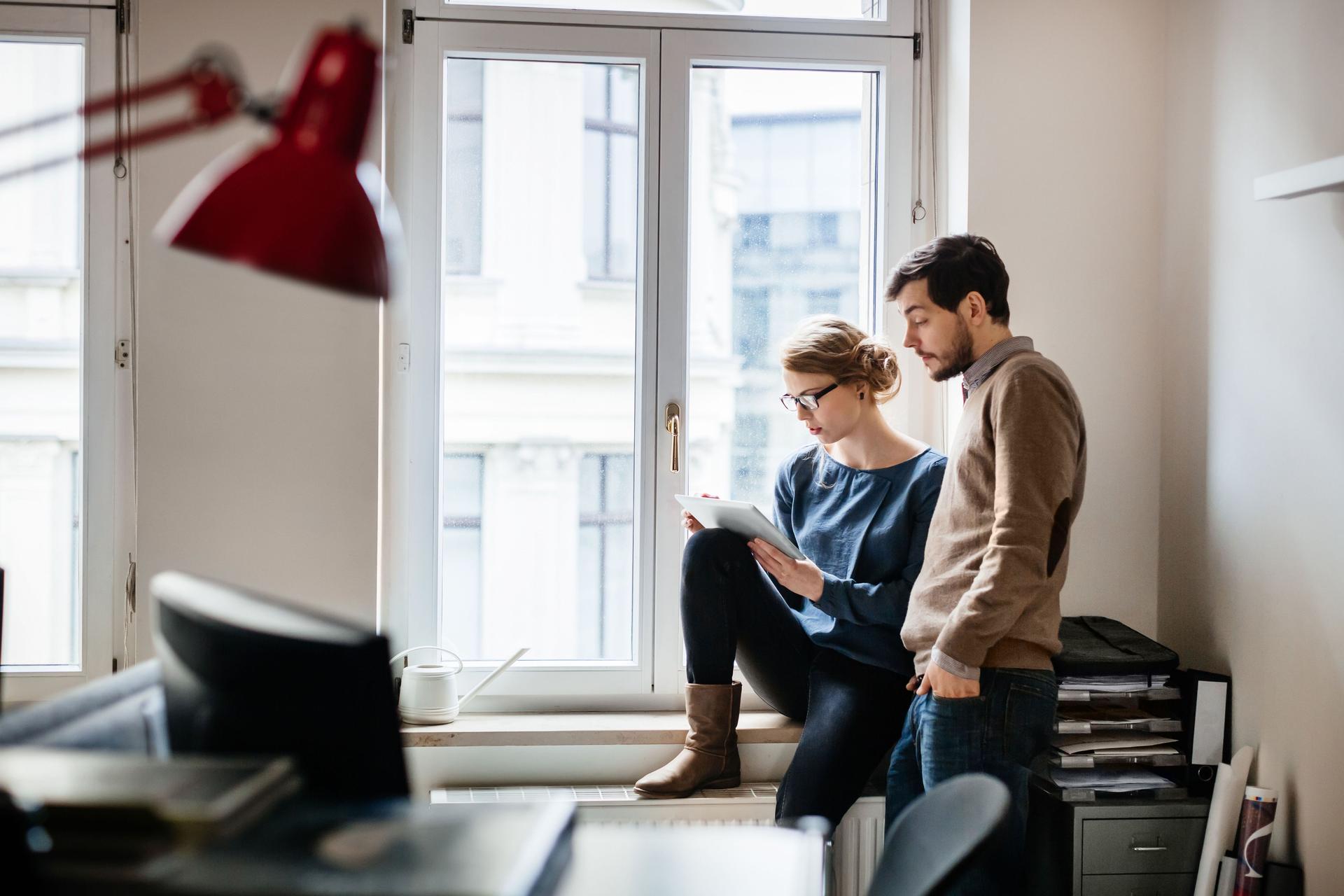 Eine junge Frau sitzt an einem Fenster in einem Büro und neben ihr steht ein junger Mann. Beide schauen auf ein digitales Tablet.