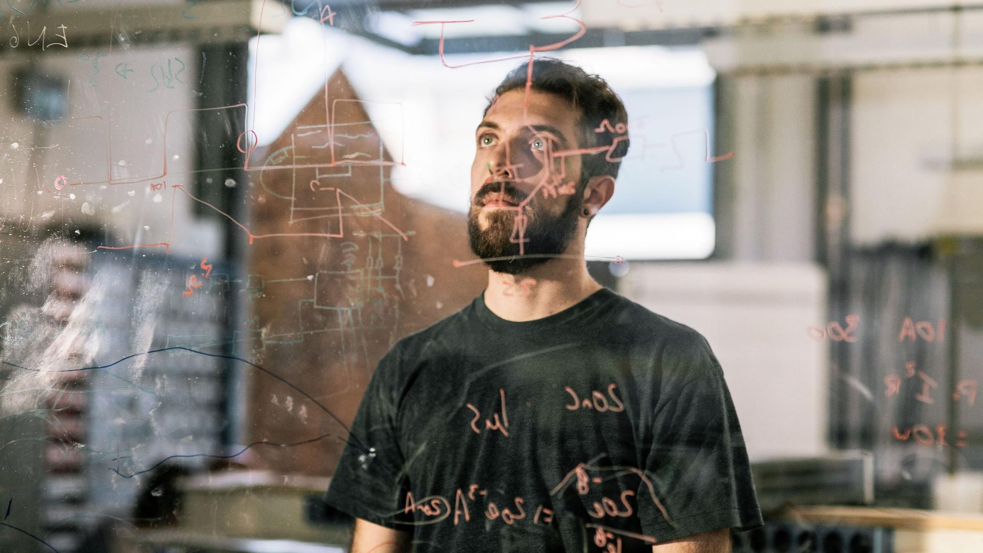 Junger Mann schaut konzentriert auf einen auf Glas gezeichneten elektronischen Schaltplan.