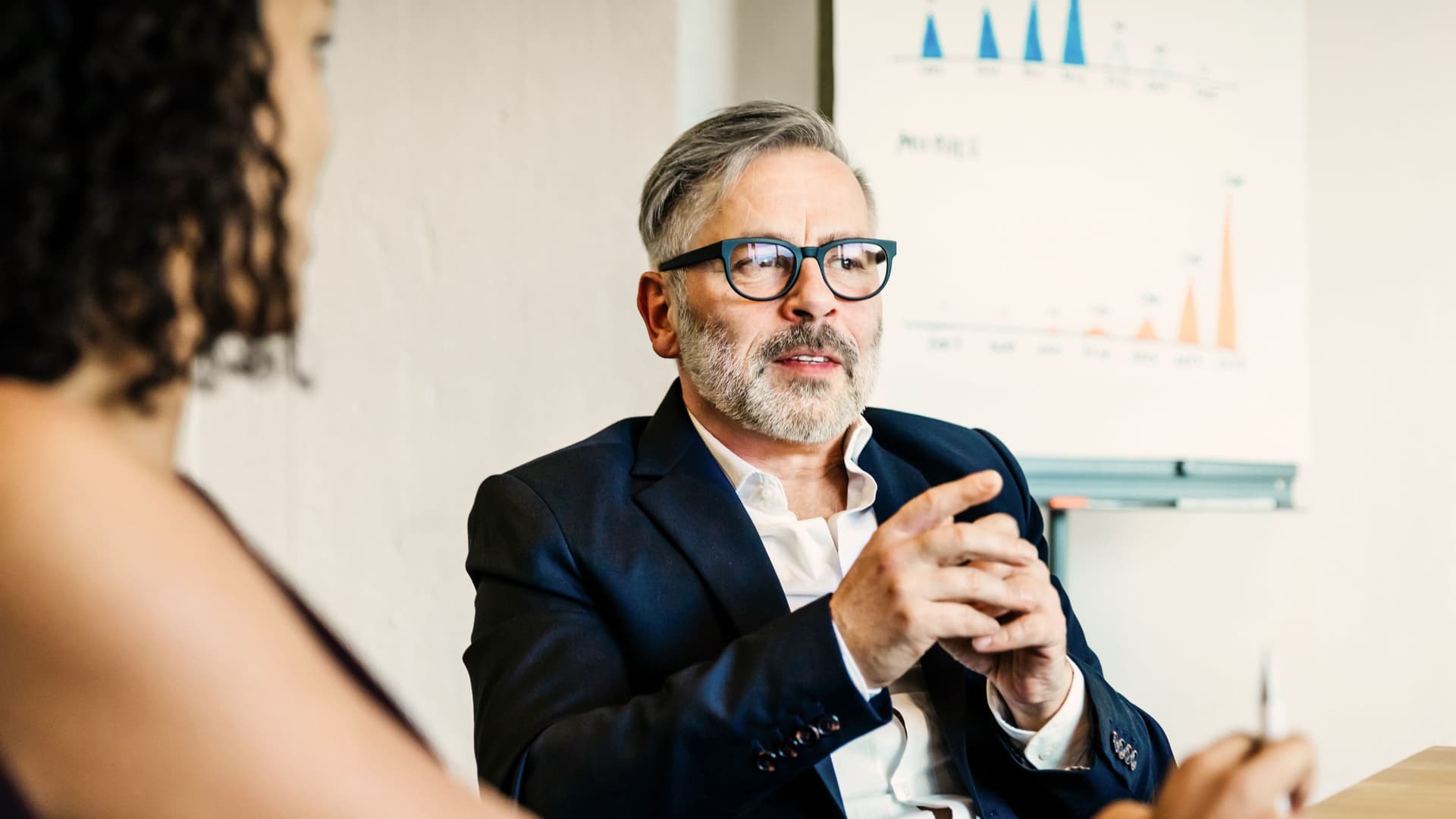 Geschäftsmann spricht während eines Meetings. Er sitzt an einem Tisch und im Hintergrund ist ein Whiteboard mit Diagrammen.