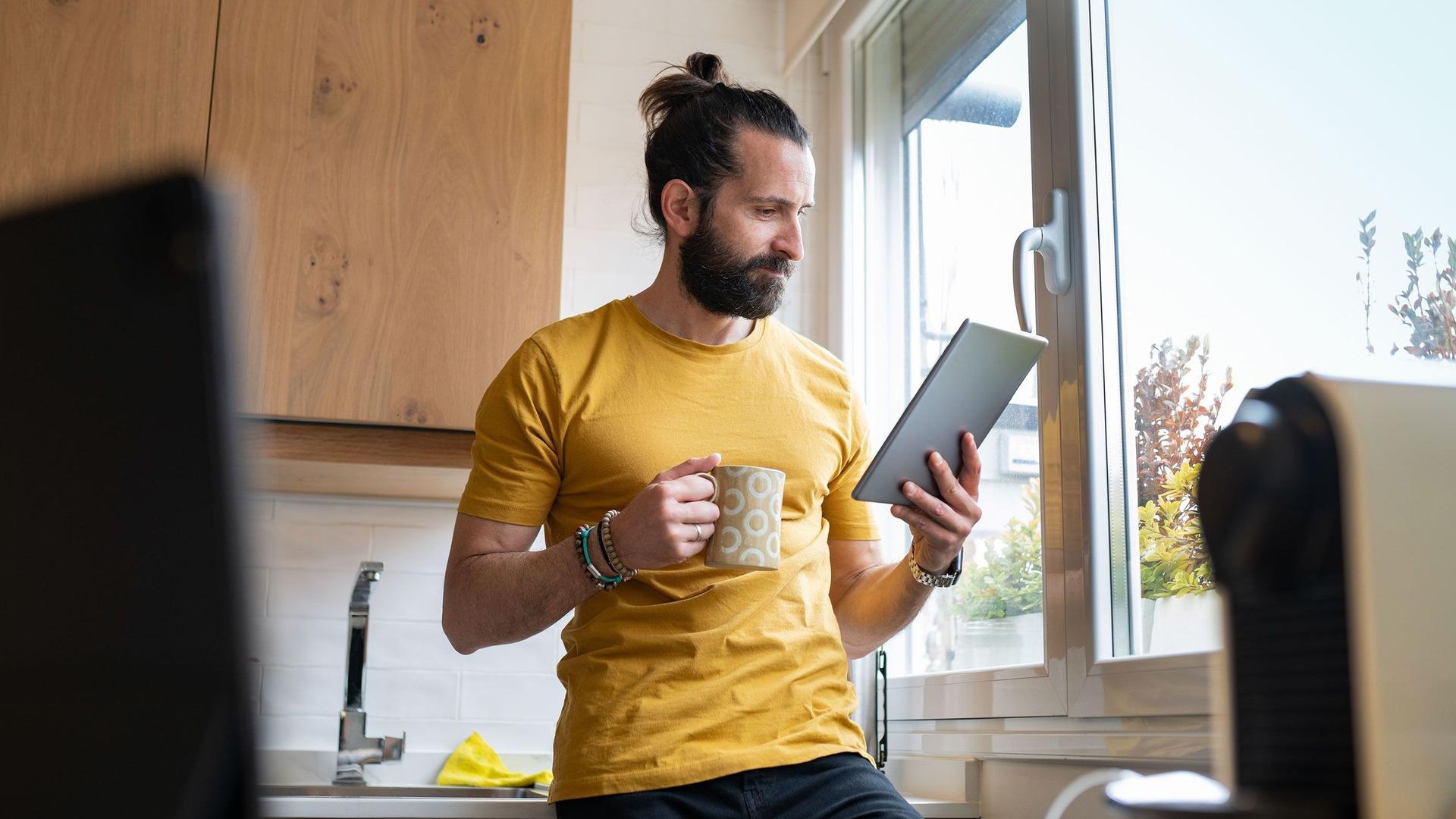 Ein junger Mann lehnt mit einem Kaffeebecher an einer Küchenzeile neben dem Fenster. Er schaut konzentriert auf ein Tablet in seiner Hand.