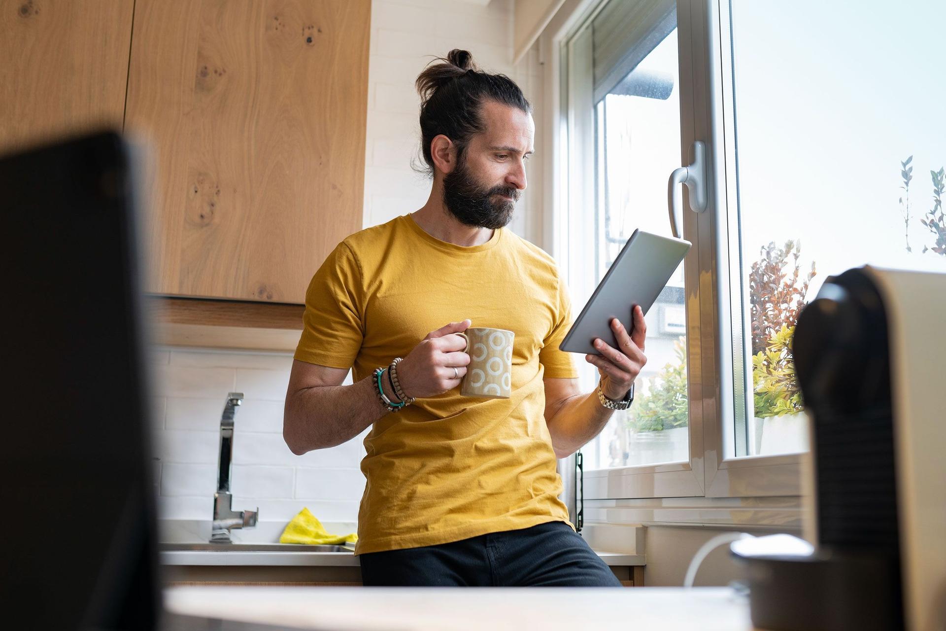 Ein junger Mann lehnt mit einem Kaffeebecher an einer Küchenzeile neben dem Fenster. Er schaut konzentriert auf ein Tablet in seiner Hand.