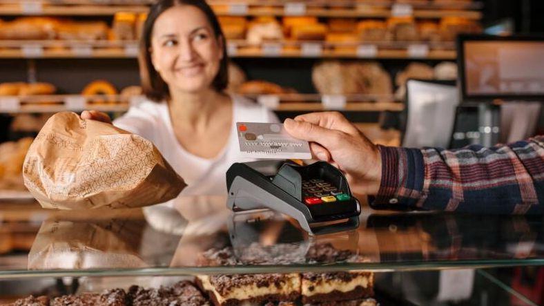 Eine männliche Hand hält in einer Bäckerei seine Sparkassenbankkarte an ein Kartenlesegerät. Im Hintergrund sieht man eine Bäckereifachangestellte die eine Brötchentüte neben ihm ablegt.