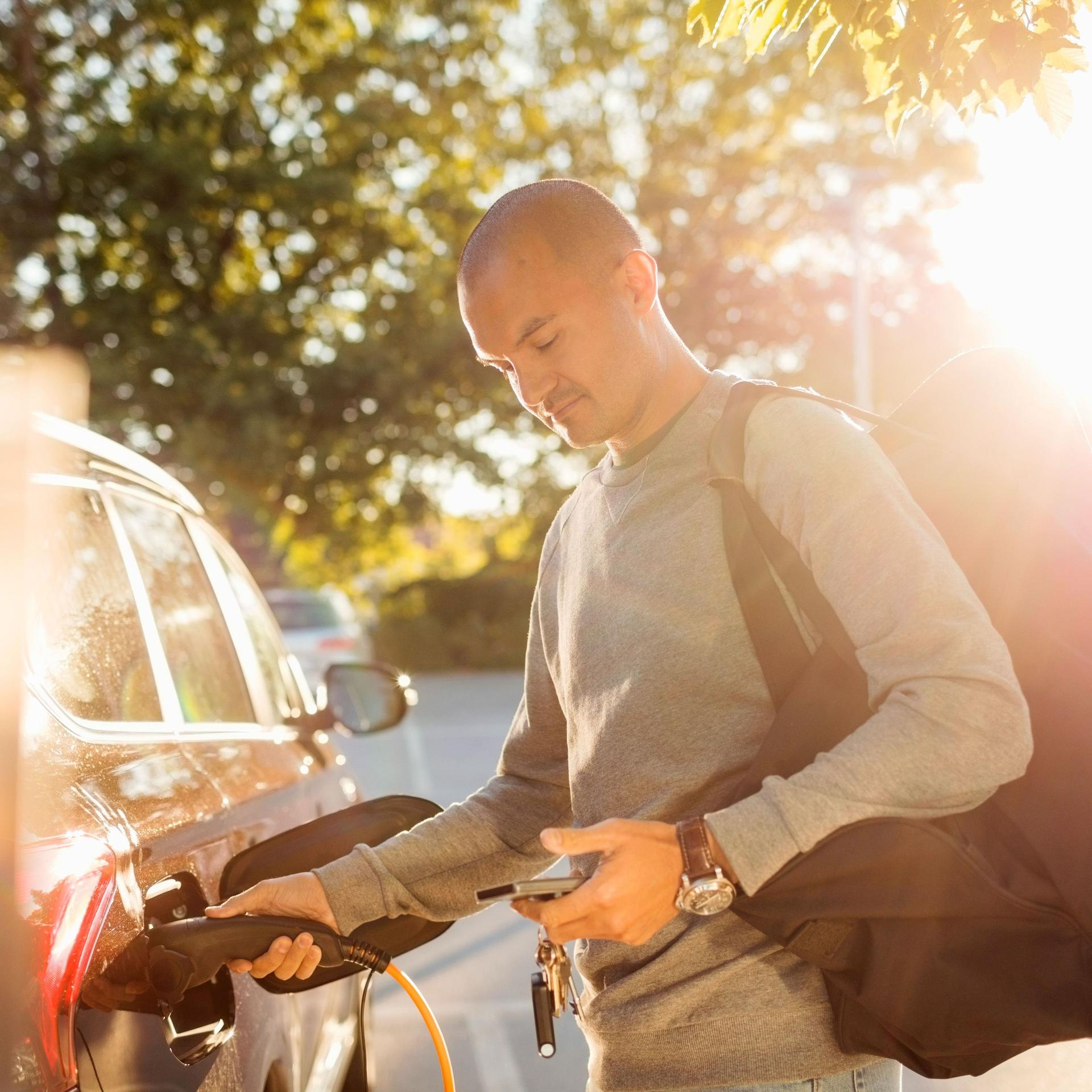 Ein Mann lädt bei Sonnenschein ein Elektroauto auf . Dabei schaut er in sein Smartphone.
