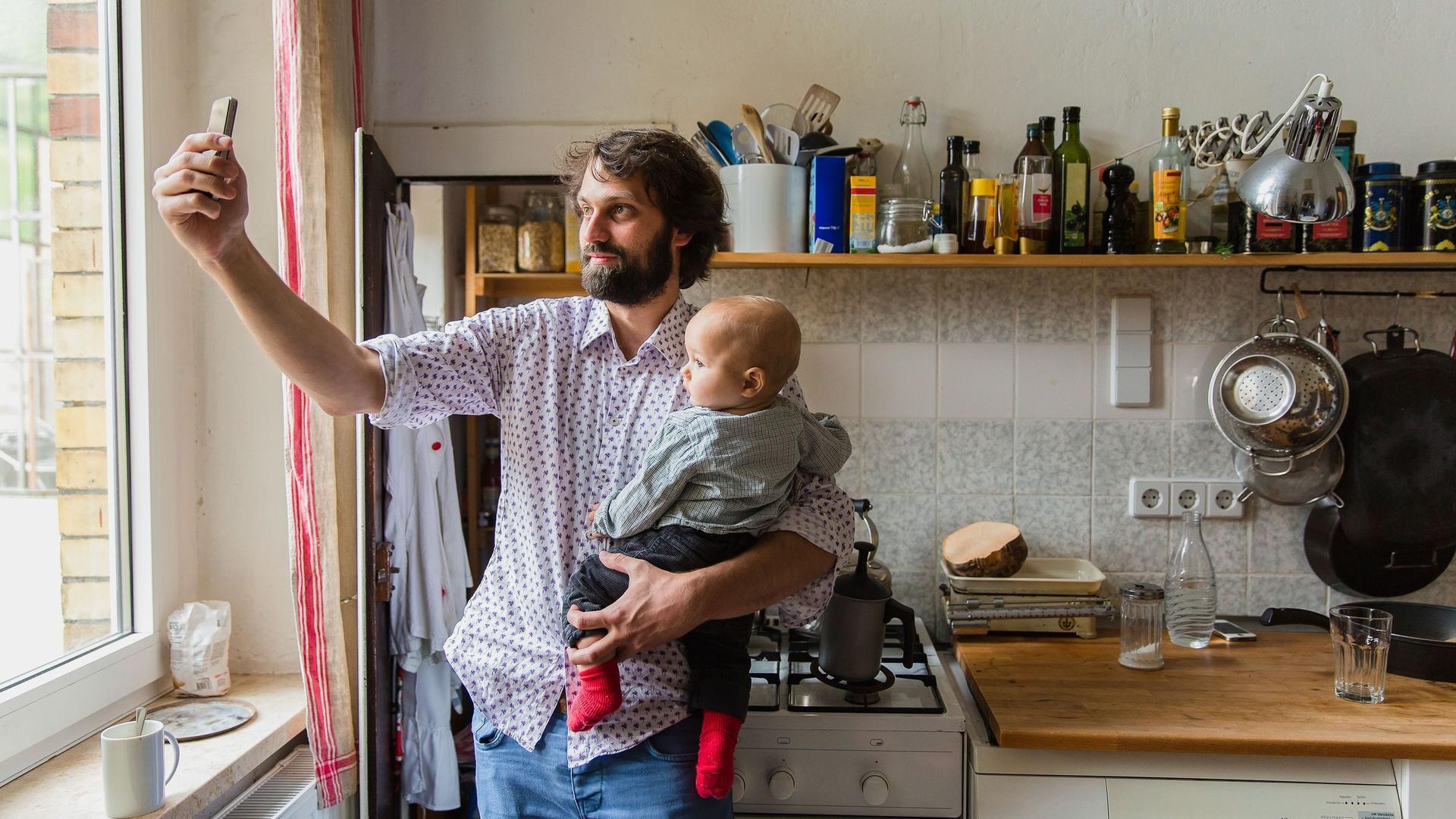 Ein junger Vater mit Baby auf dem Arm steht in einer Küche und macht ein Selfie von sich und seinem Kind.