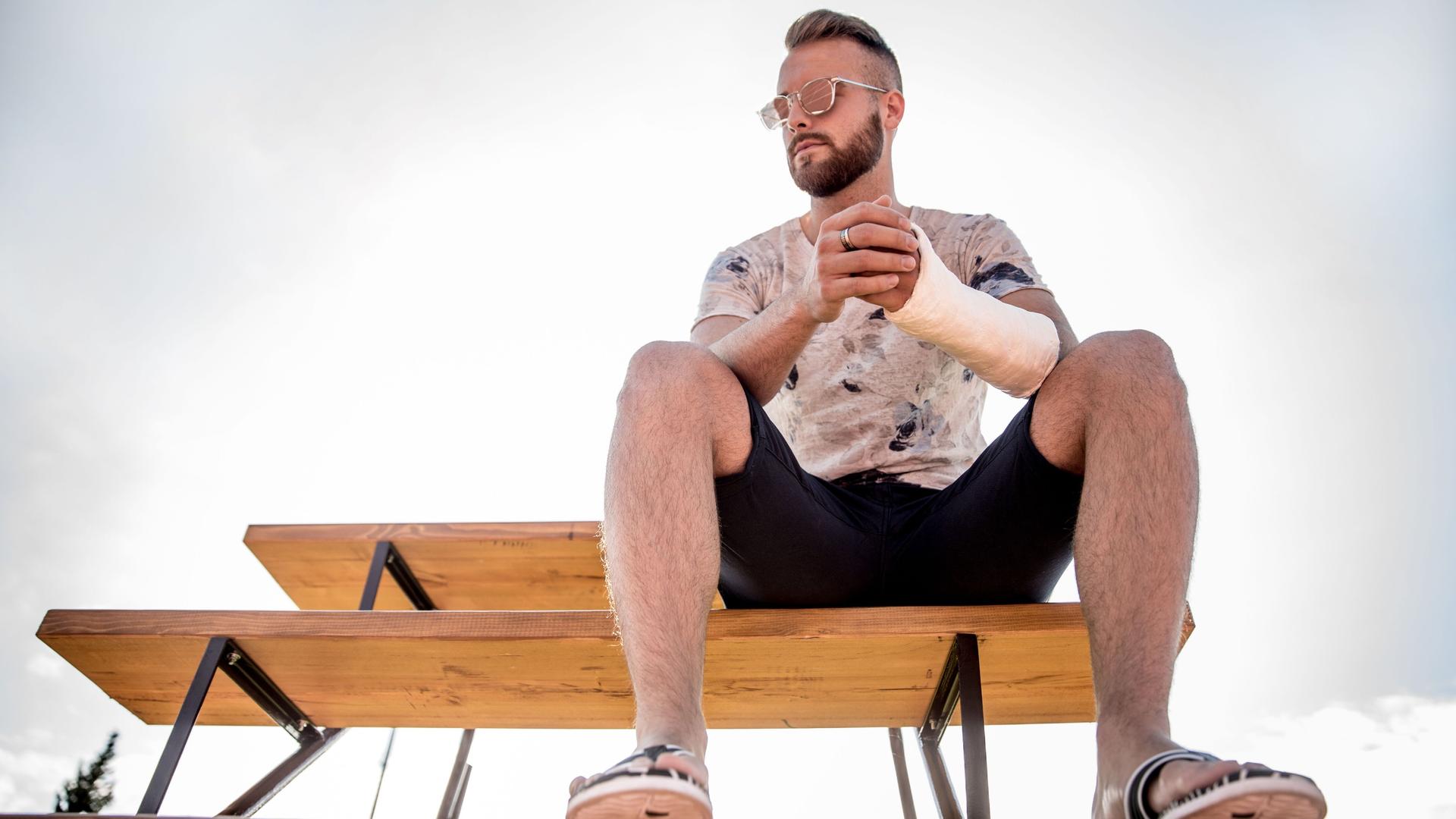 Ein Mann mit Sonnenbrille und gebrochenem Arm sitzt auf einer Bank in der Sonne.