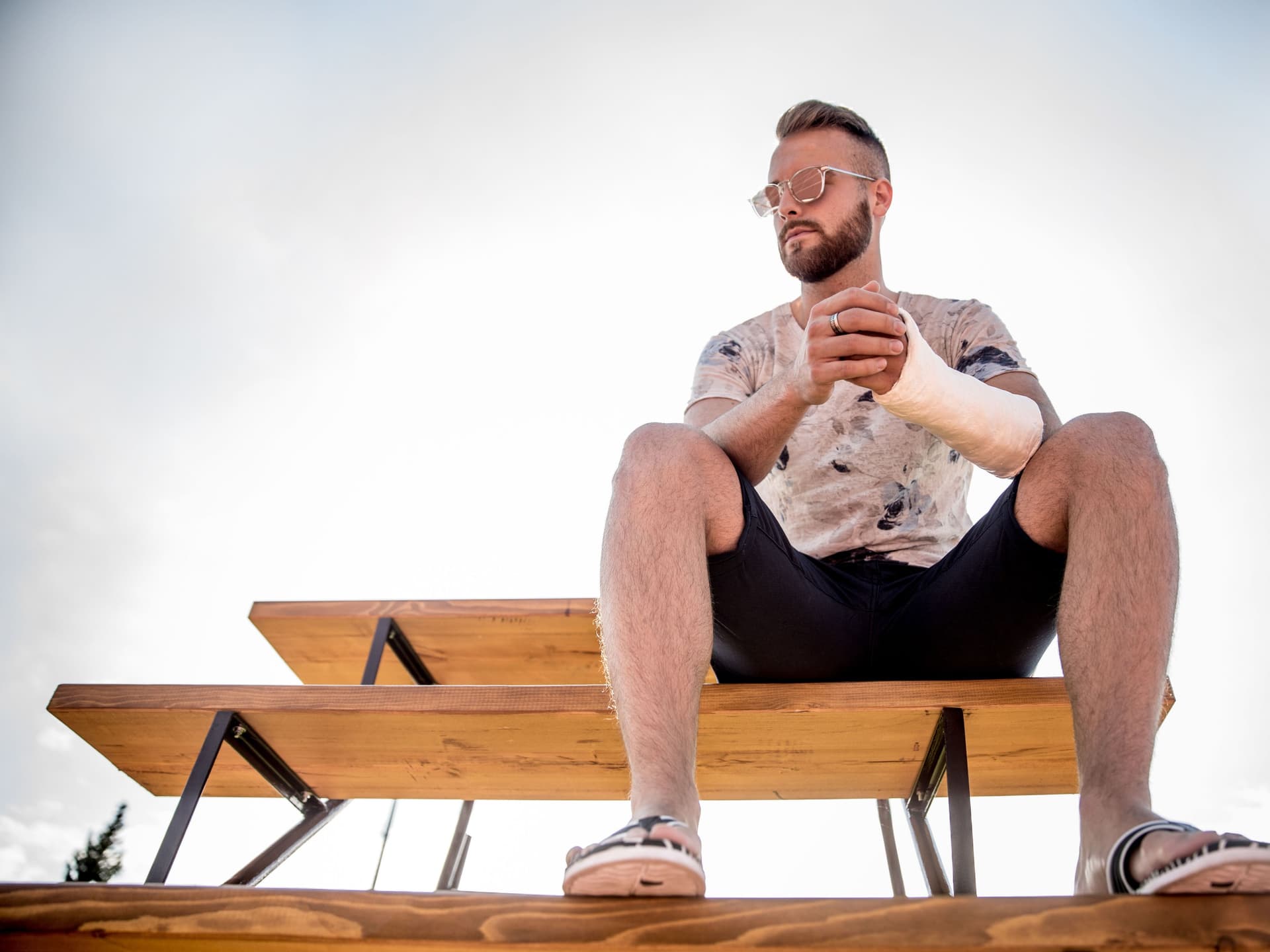 Ein Mann mit Sonnenbrille und gebrochenem Arm sitzt auf einer Bank in der Sonne.