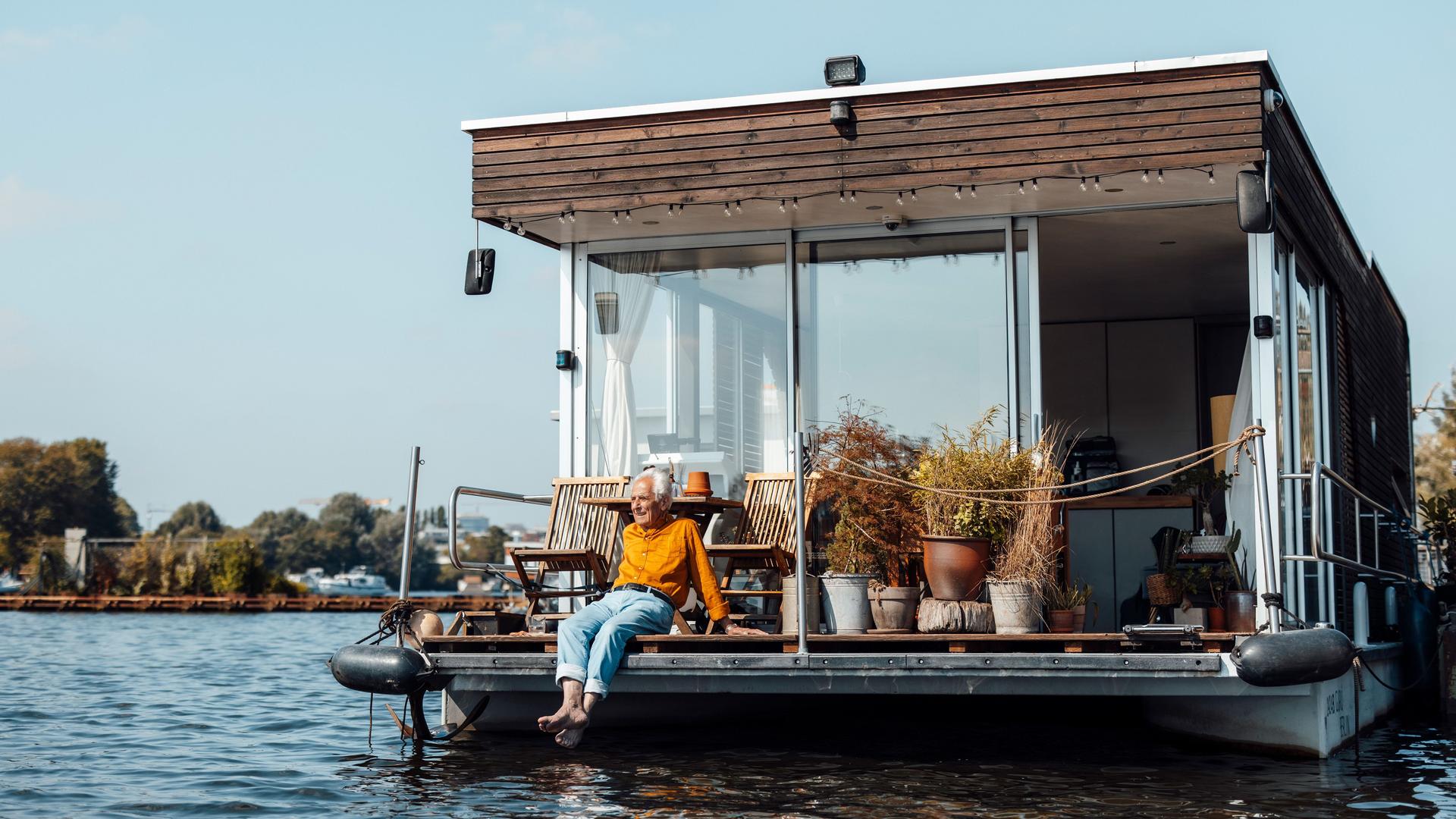 Mann mit gruaen Haaren und einem gelben Pulli sitzt am Heck eines Hausboots und lässt die Beine baumeln.