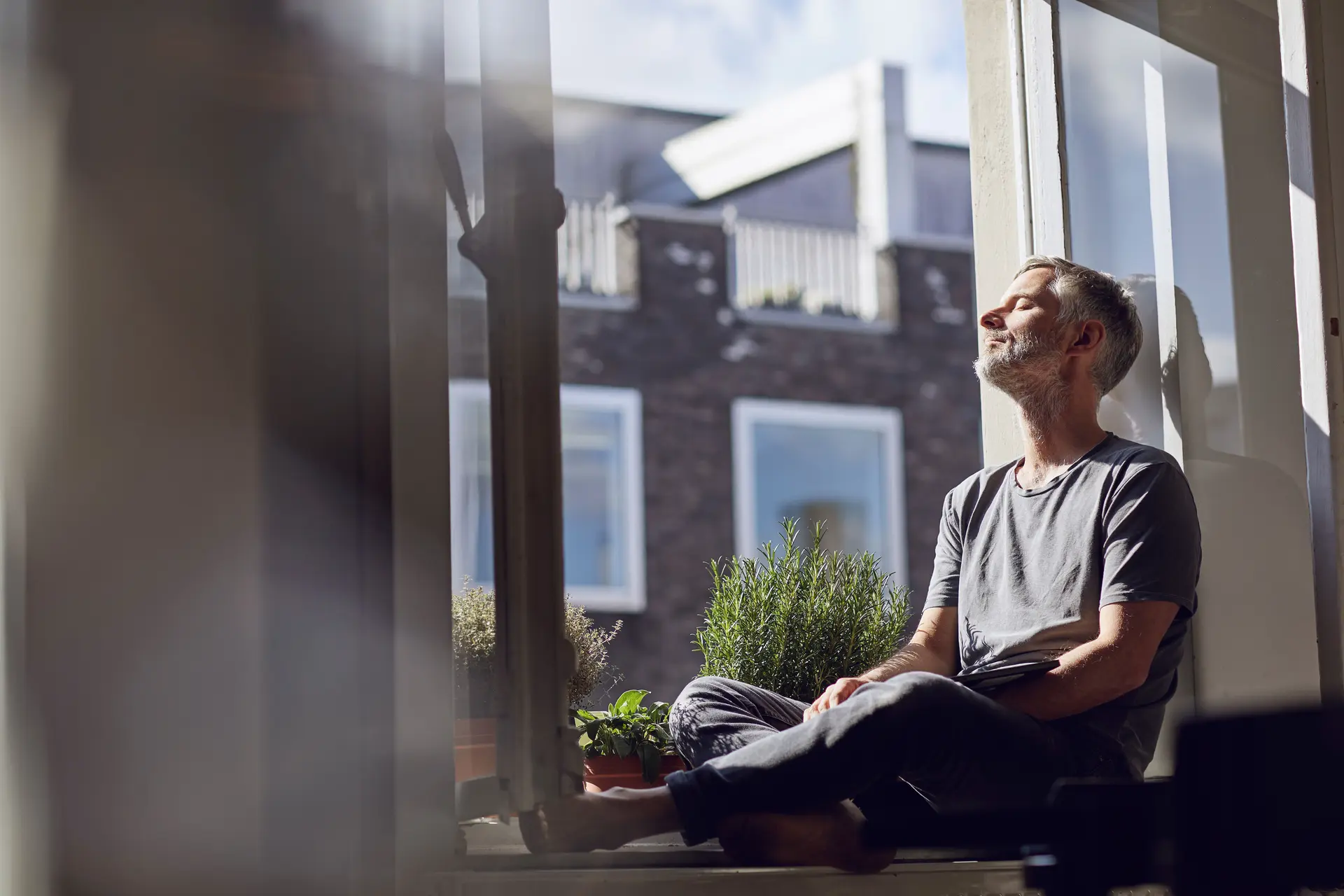 Ein Mann mit grauem Bart sitzt mit geschlossenen Augen auf dem Fenstersims und genießt die Sonne.