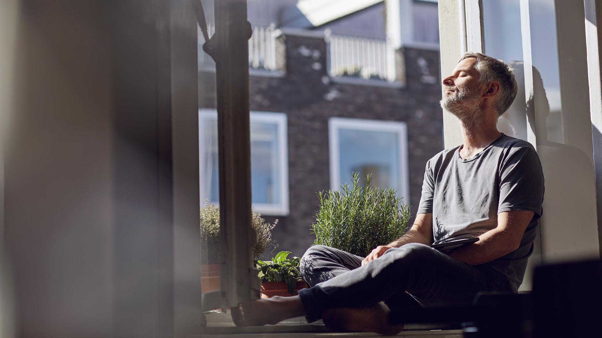 Ein Mann mit grauem Bart sitzt mit geschlossenen Augen auf dem Fenstersims und genießt die Sonne.