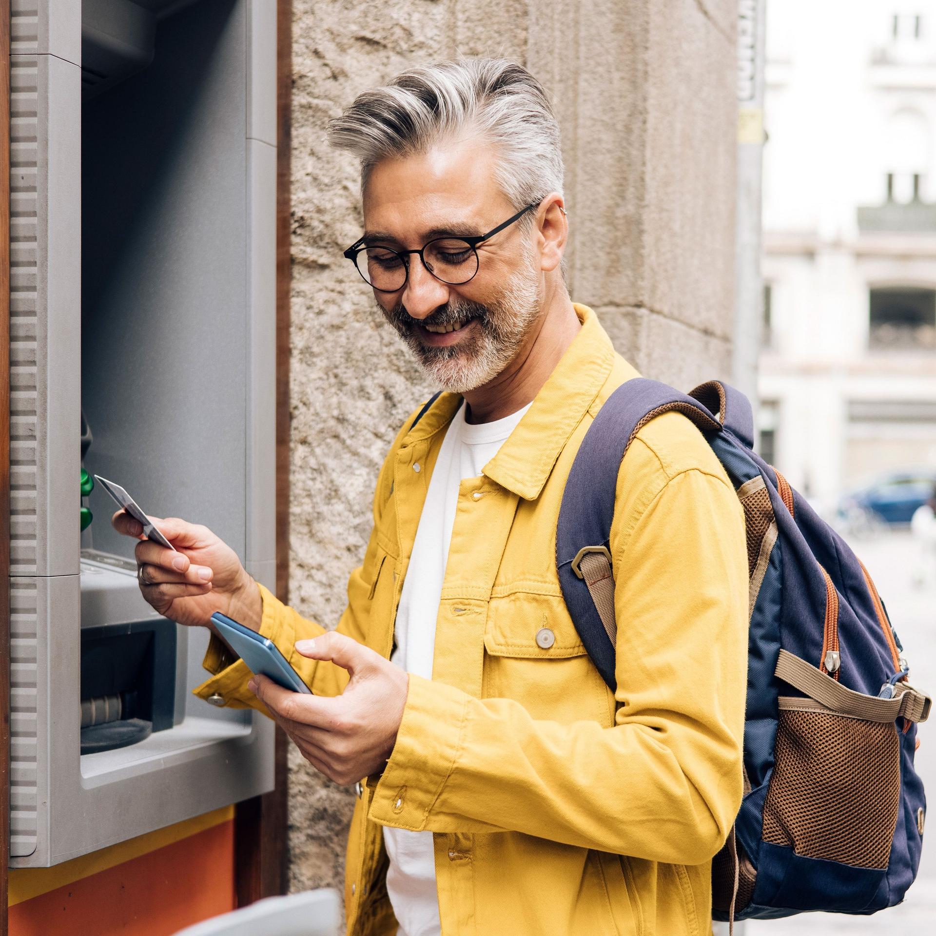 Mann mit grauen Haaren, Brille und gelber Jacke steht lächelnd vor einem Geldautomat. Er schaut auf sein Handy.