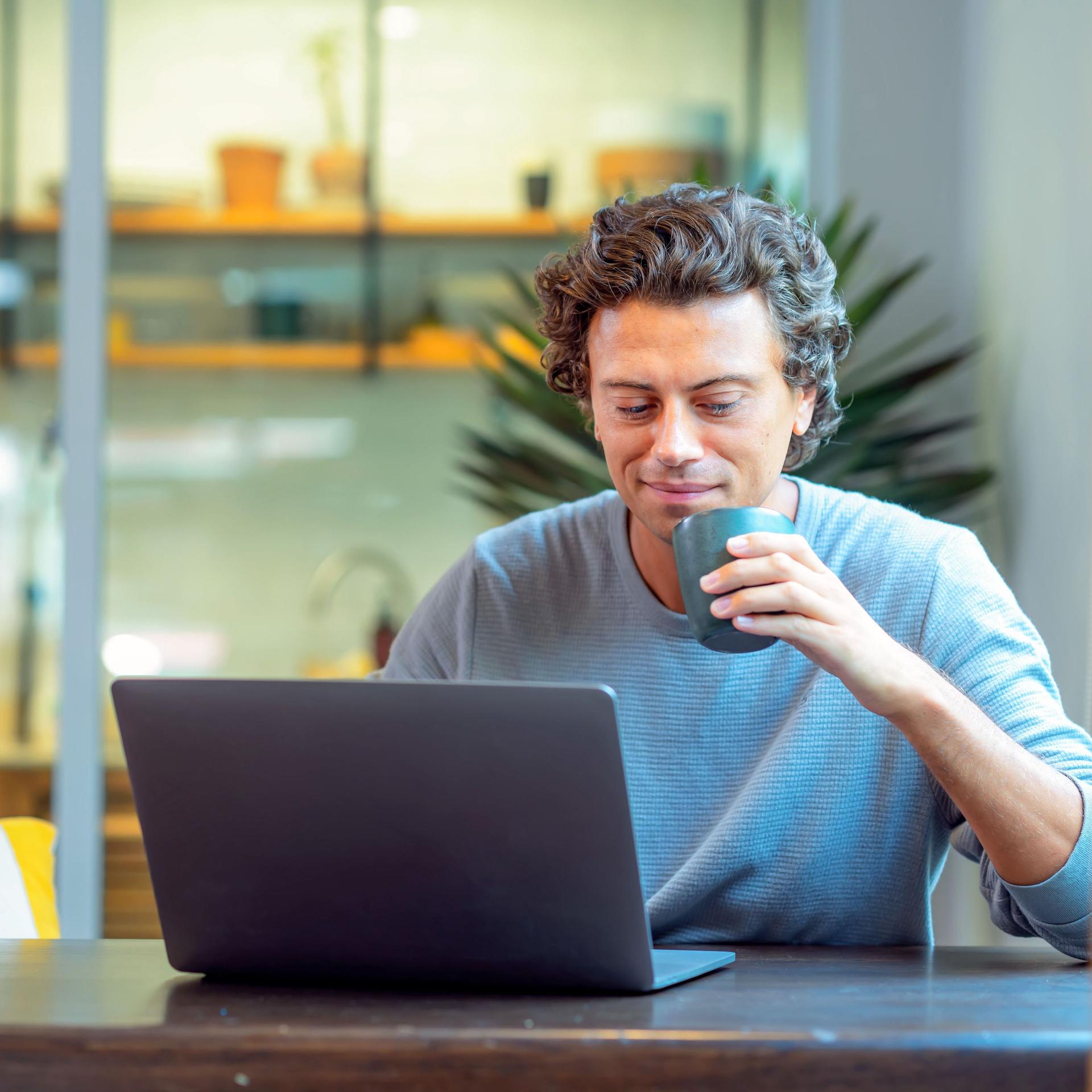 Ein junger Mann sitzt lächelnd und mit Kaffeetasse in der Hand an einem Laptop Computer.