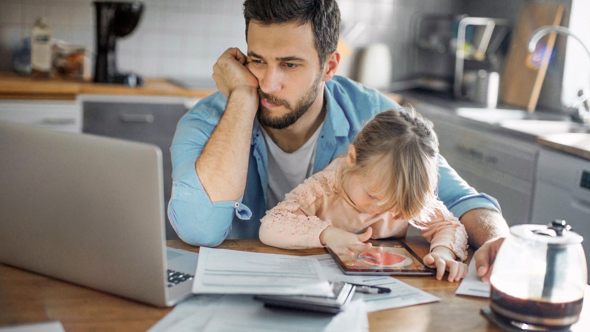 Missmutig schauender Vater mit seiner kleinen Tochter auf dem Schoß schaut in einen Laptop. Er sitzt in seiner Küche und vor ihm liegen Unterlagen.
