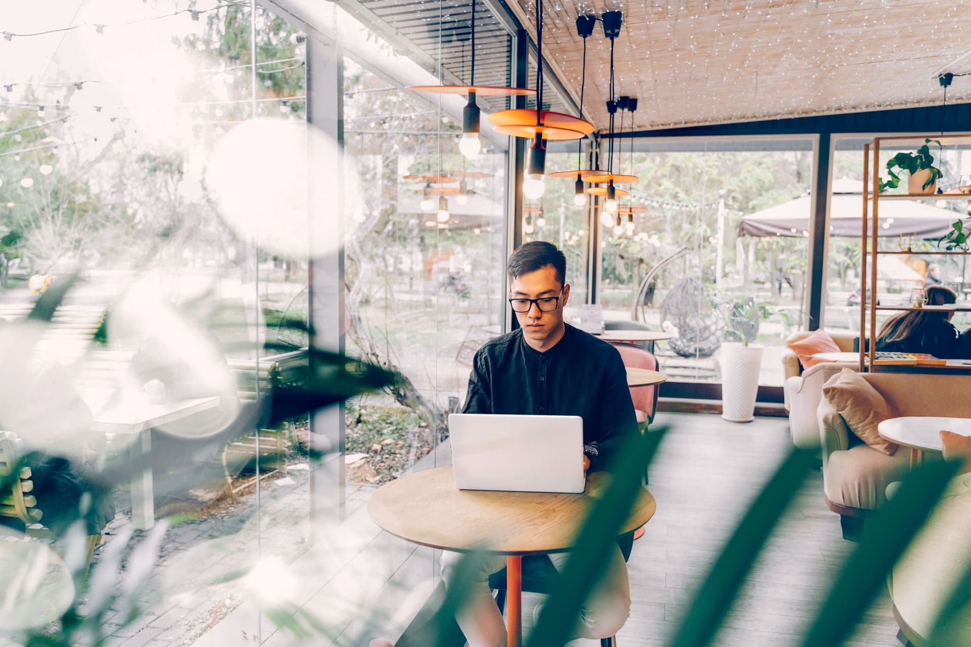 Mann mit Laptop sitzt an einem kleinen Tisch in einem Cafe vor einer großen Glasfront.