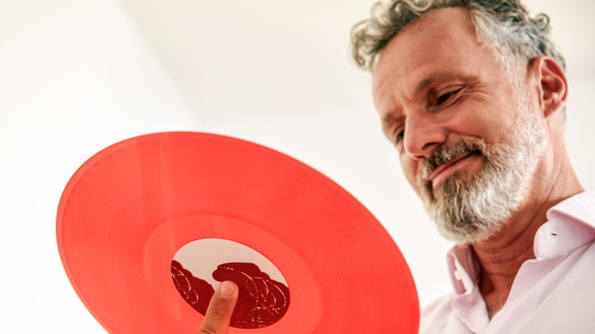 Mann im mittleren Alter lächelt und schaut auf eine rote Schallplatte auf seiner Hand. Von unten aus der Froschperspektive fotografiert.
