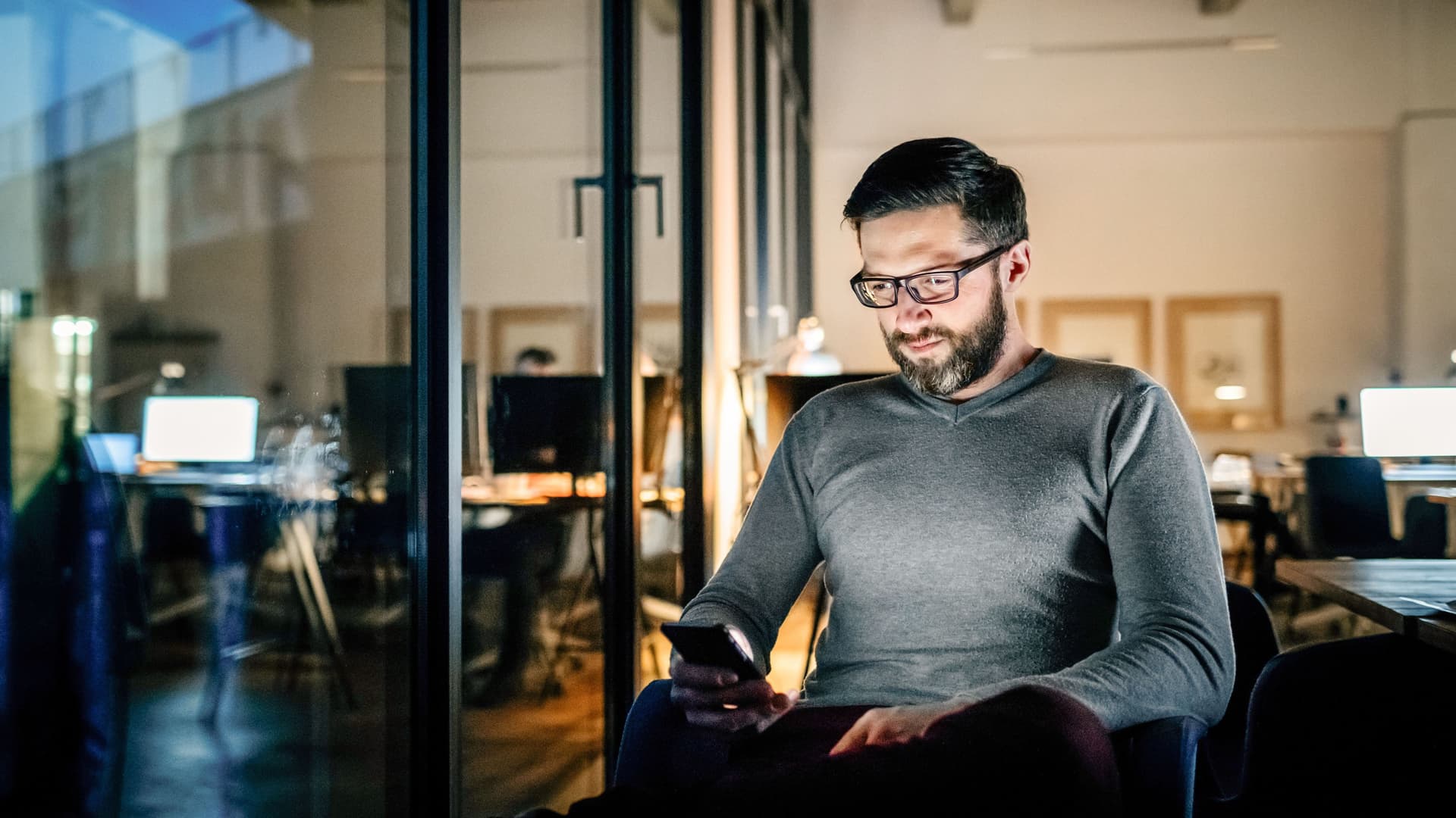Mann mit Smartphone in der Hand sitzt nachts alleine im Büro.
