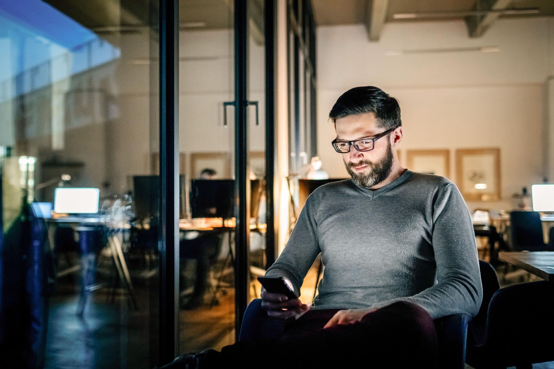 Mann mit Smartphone in der Hand sitzt nachts alleine im Büro.