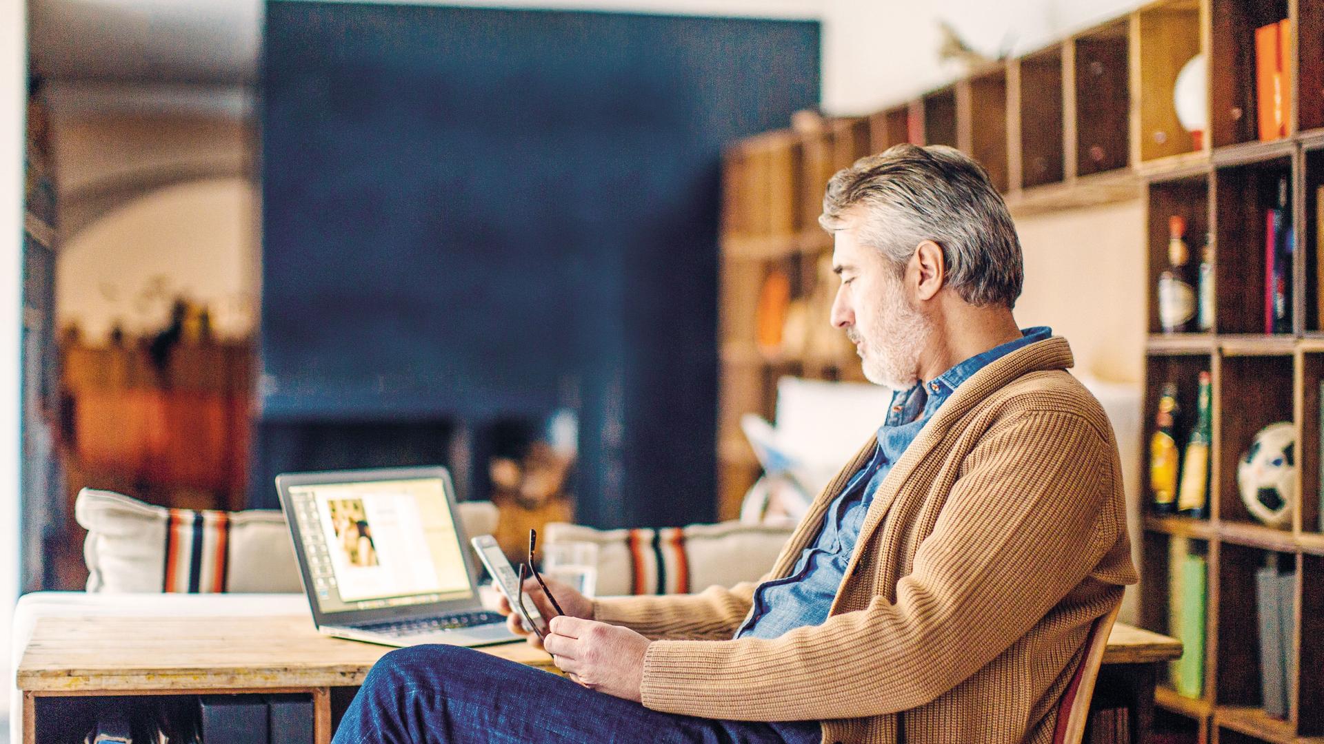 Mann mittleren Alters sitzt in einem Arbeitszimmer am Schreibtisch, benutzt sein Smartphone und hat einen Laptop vor sich stehen