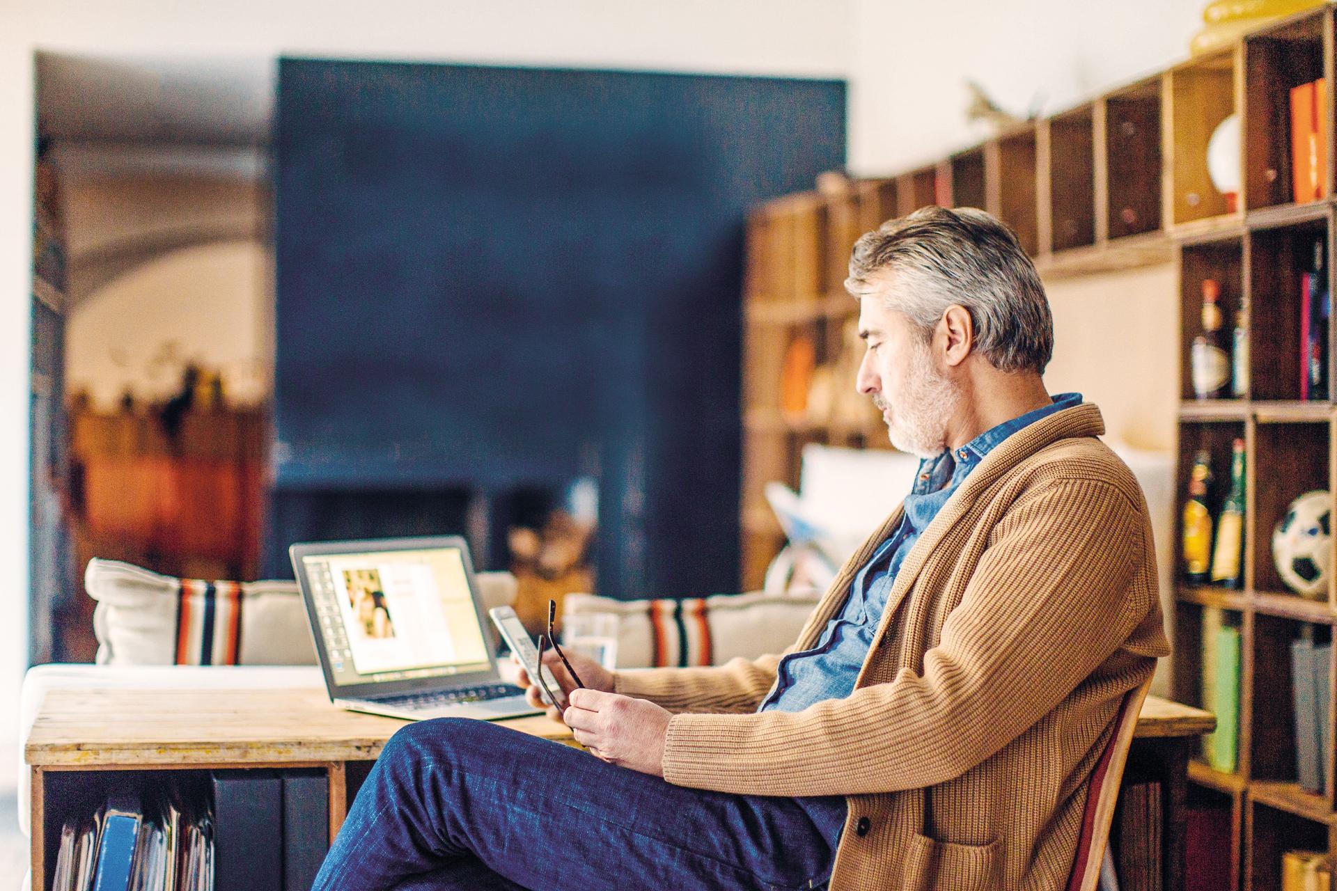 Mann mittleren Alters sitzt in einem Arbeitszimmer am Schreibtisch, benutzt sein Smartphone und hat einen Laptop vor sich stehen