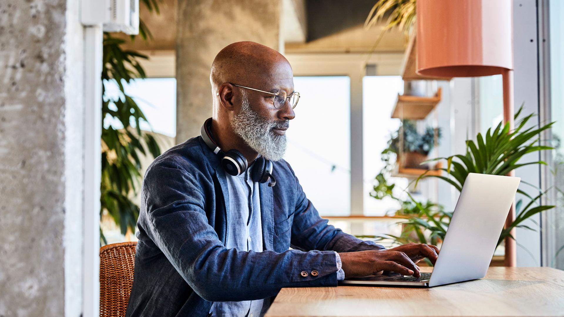 Mann mittleren Alters, der an einem Holztisch sitzt und an einem Laptop arbeitet. Im Hintergrund sind viele Pflanzen.